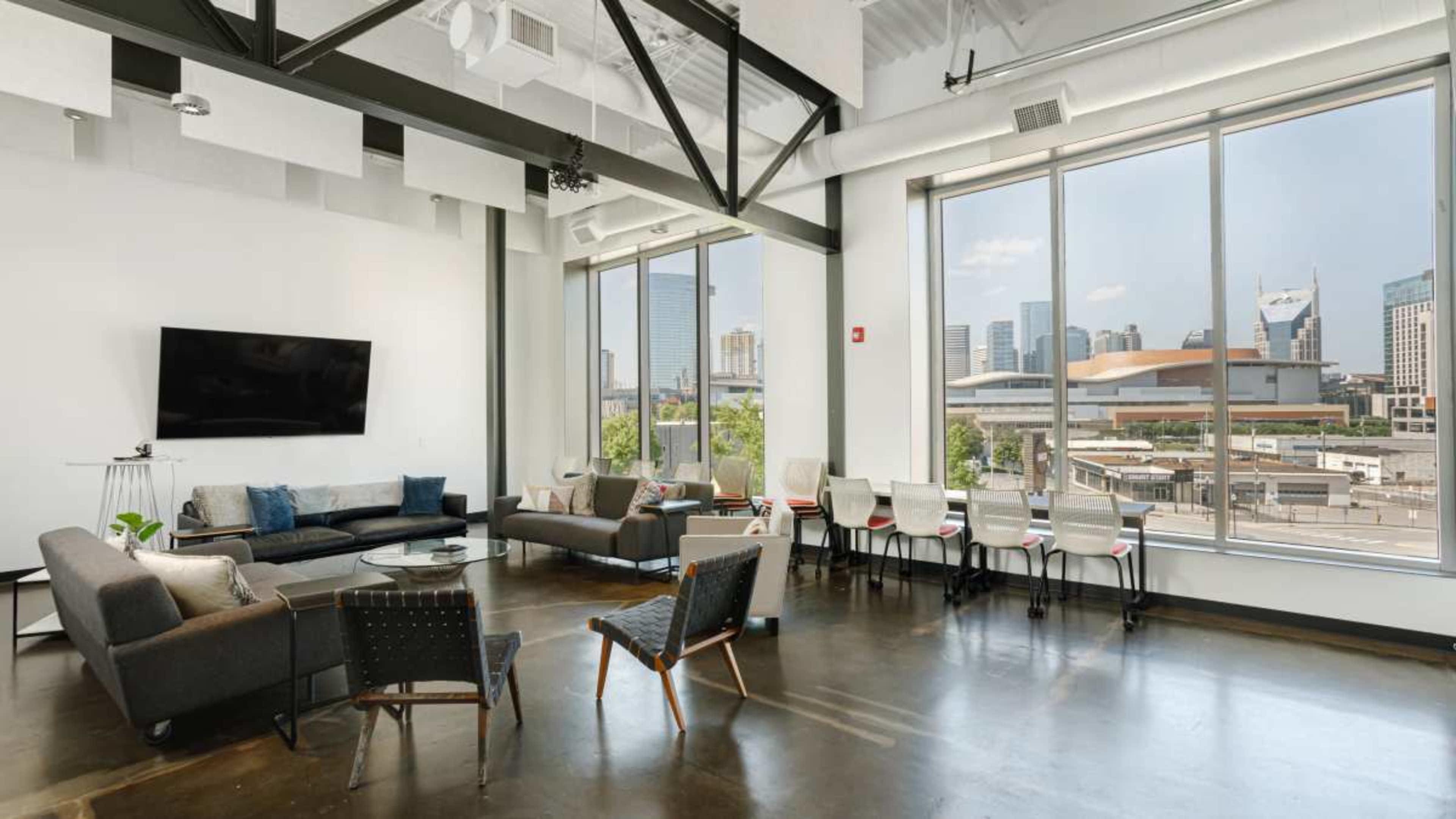 The image shows a modern office space with large windows, offering a view of city skyscrapers, and furnished with seating areas and a dining table.