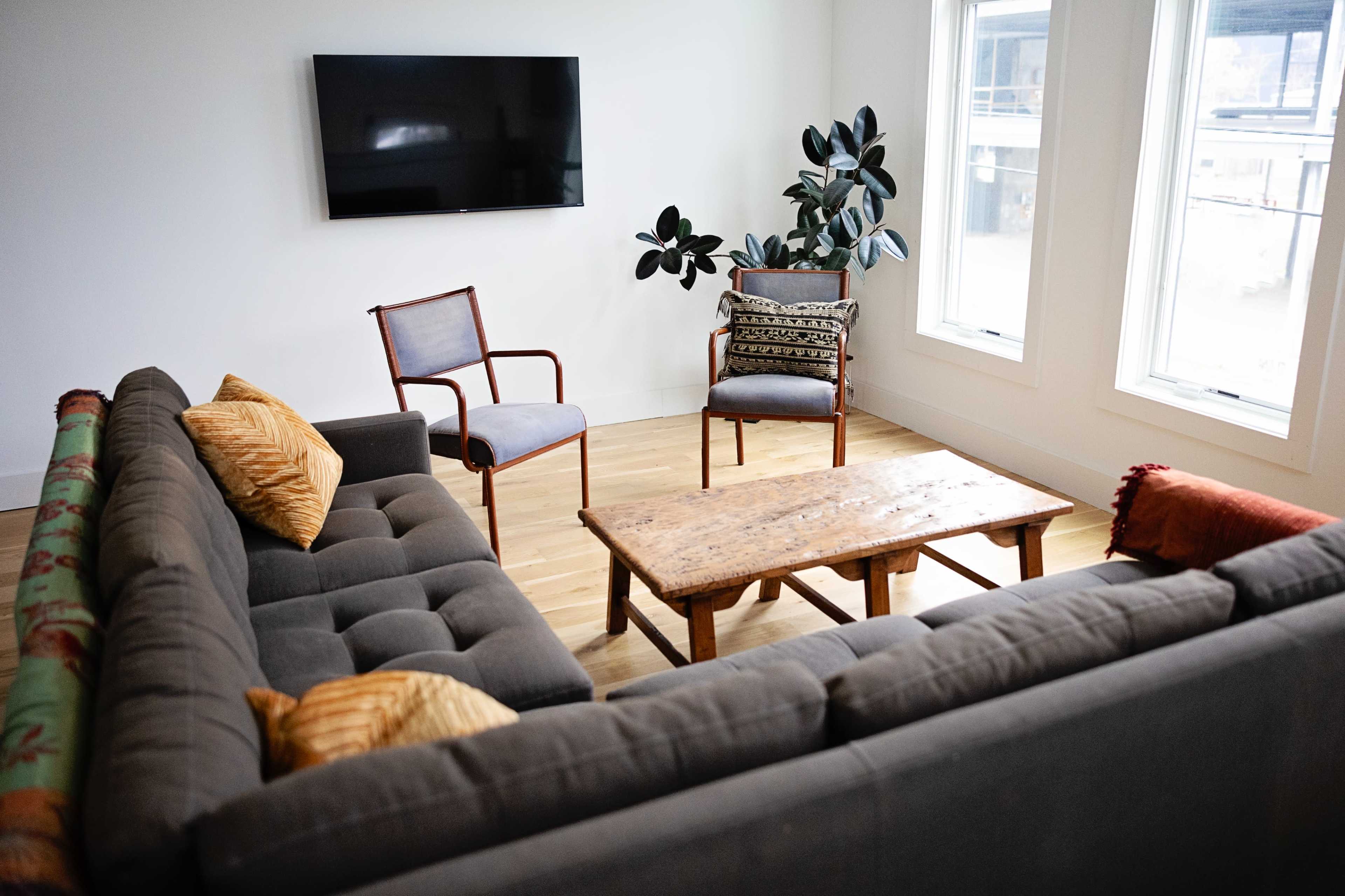 The image shows a modern living room featuring a gray sofa, two chairs, a wooden coffee table, and a large television mounted on the wall.