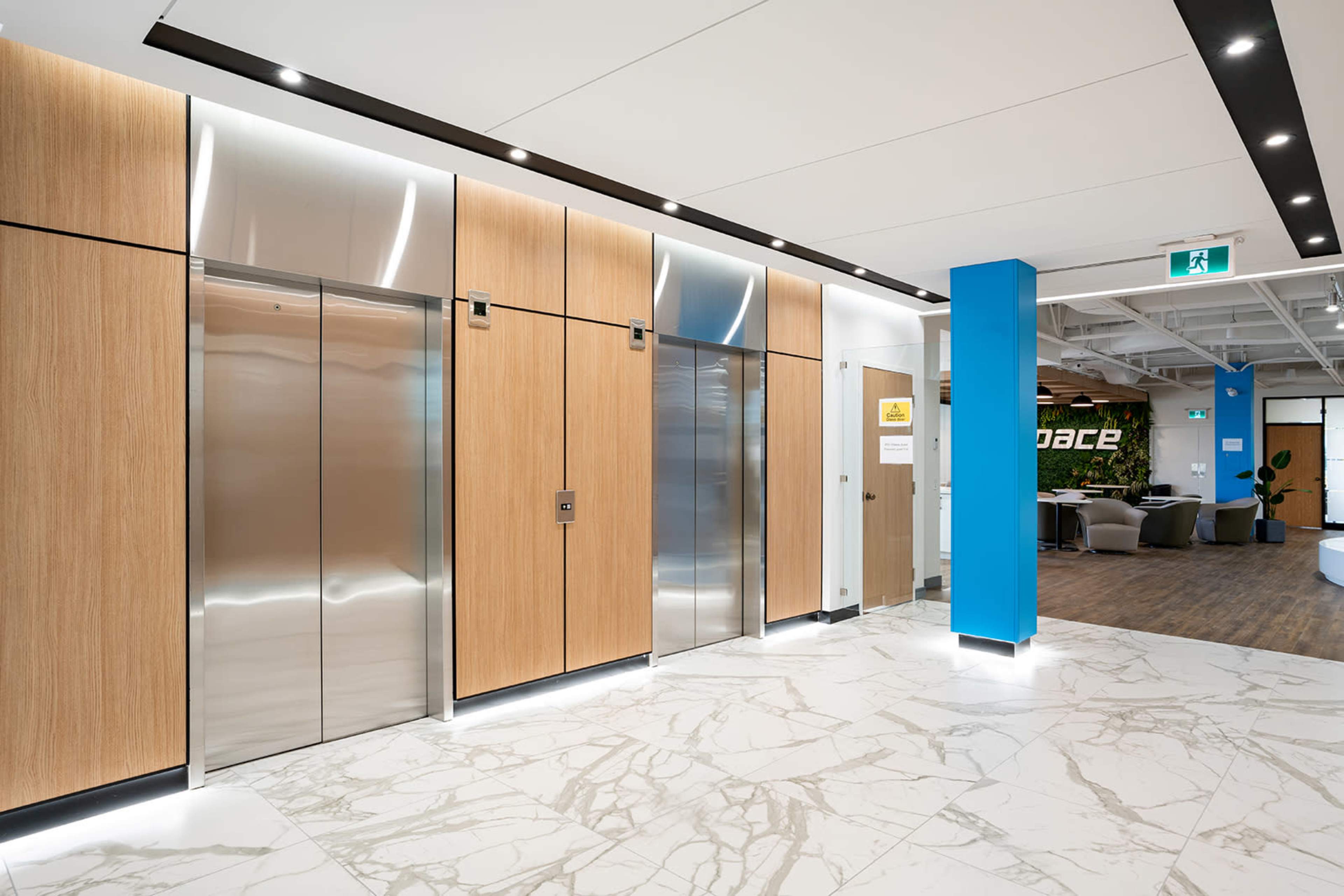 A modern lobby area with wooden elevator doors, a sleek metal elevator, and a blue wall featuring a sign, along with a marble floor and seating area in the background.