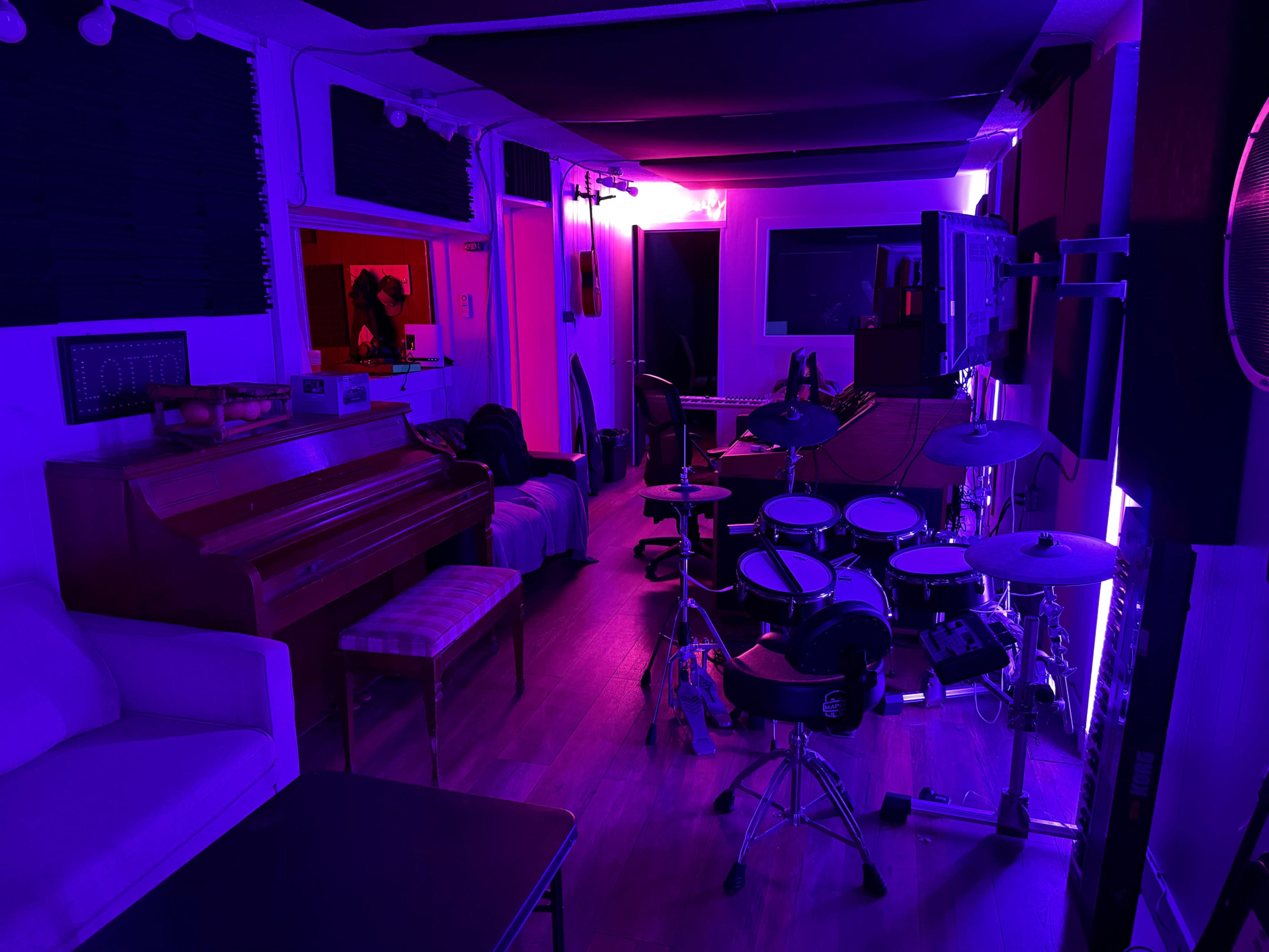 The image shows a music studio or rehearsal space with a piano, electronic drum set, and various seating areas, illuminated by purple and red lighting.