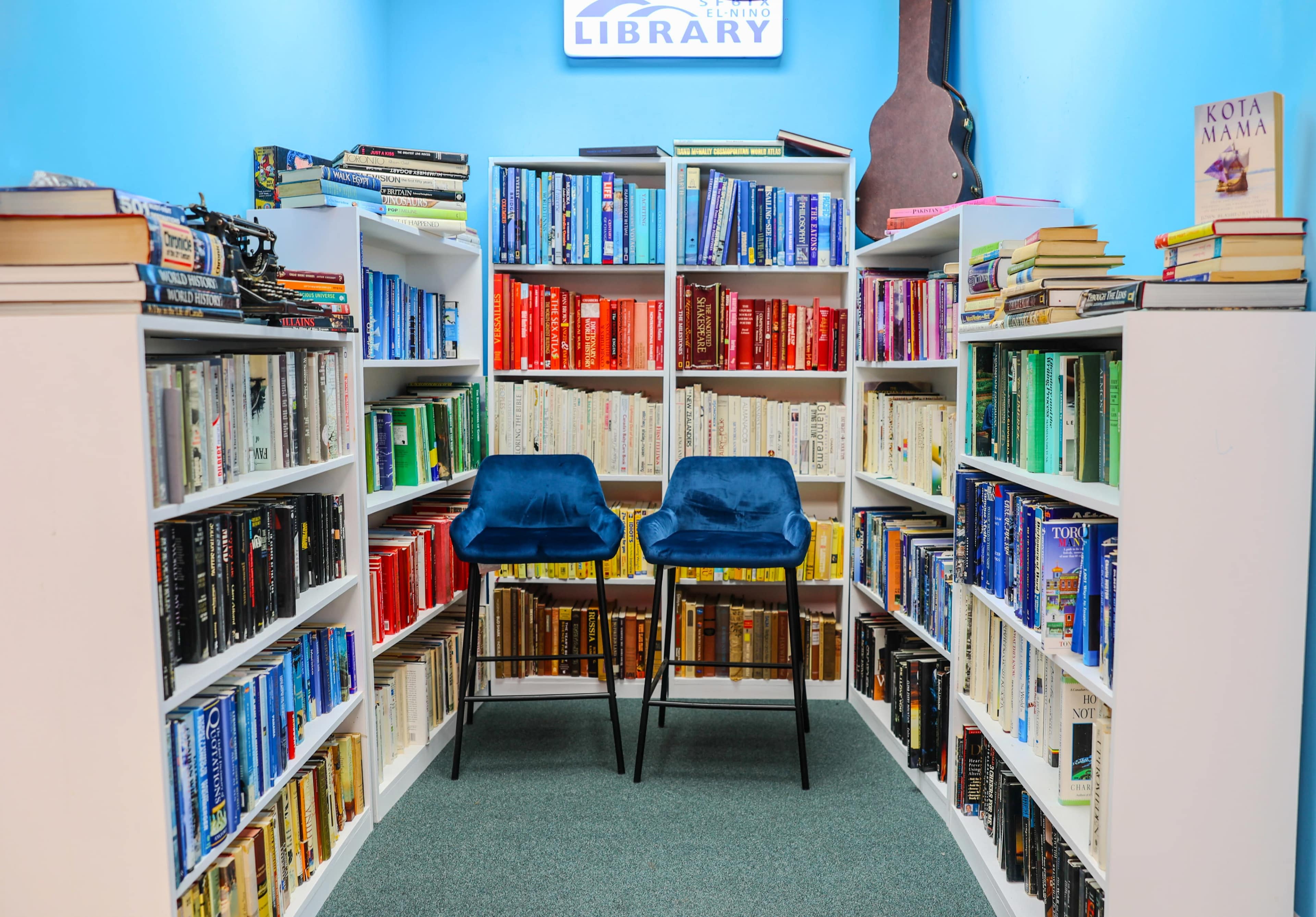 The image shows a small library with white bookshelves filled with books arranged by color, flanking two blue velvet chairs in the center against a blue wall.