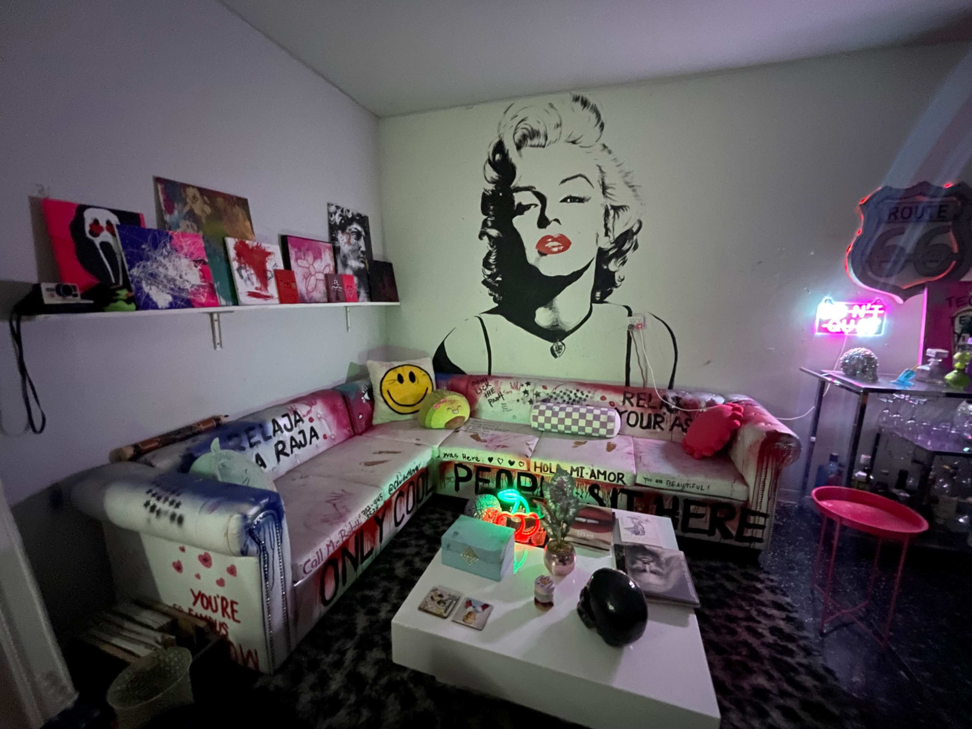 The image shows a colorful room featuring a large couch with graffiti-style art, a wall mural of a woman, and various decor items, including smiley face cushions and neon lights.