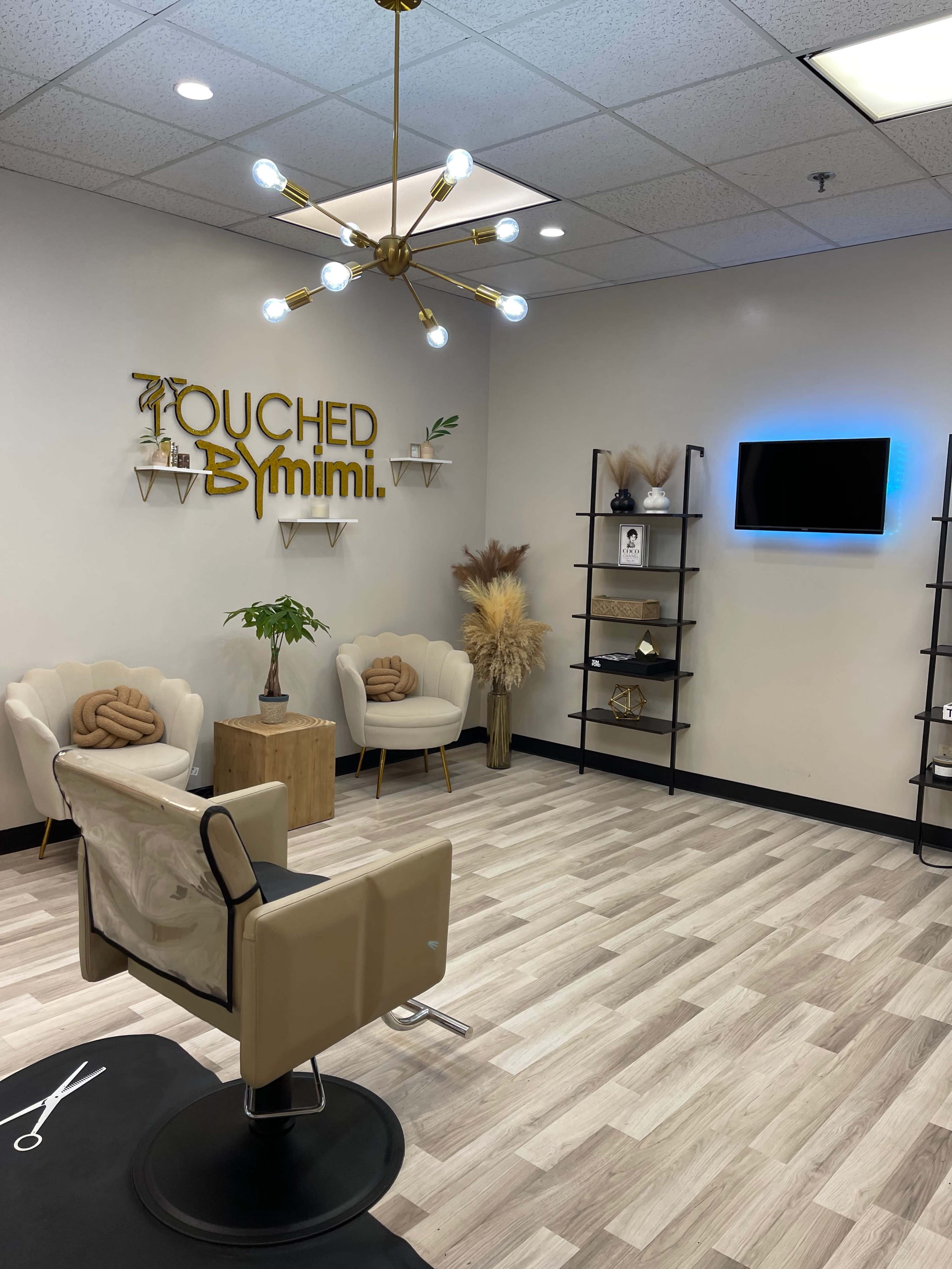 The image shows a salon interior with two white chairs, a wooden table, decorative plants, and a wall sign that reads "Touched By Mimi" in gold lettering.