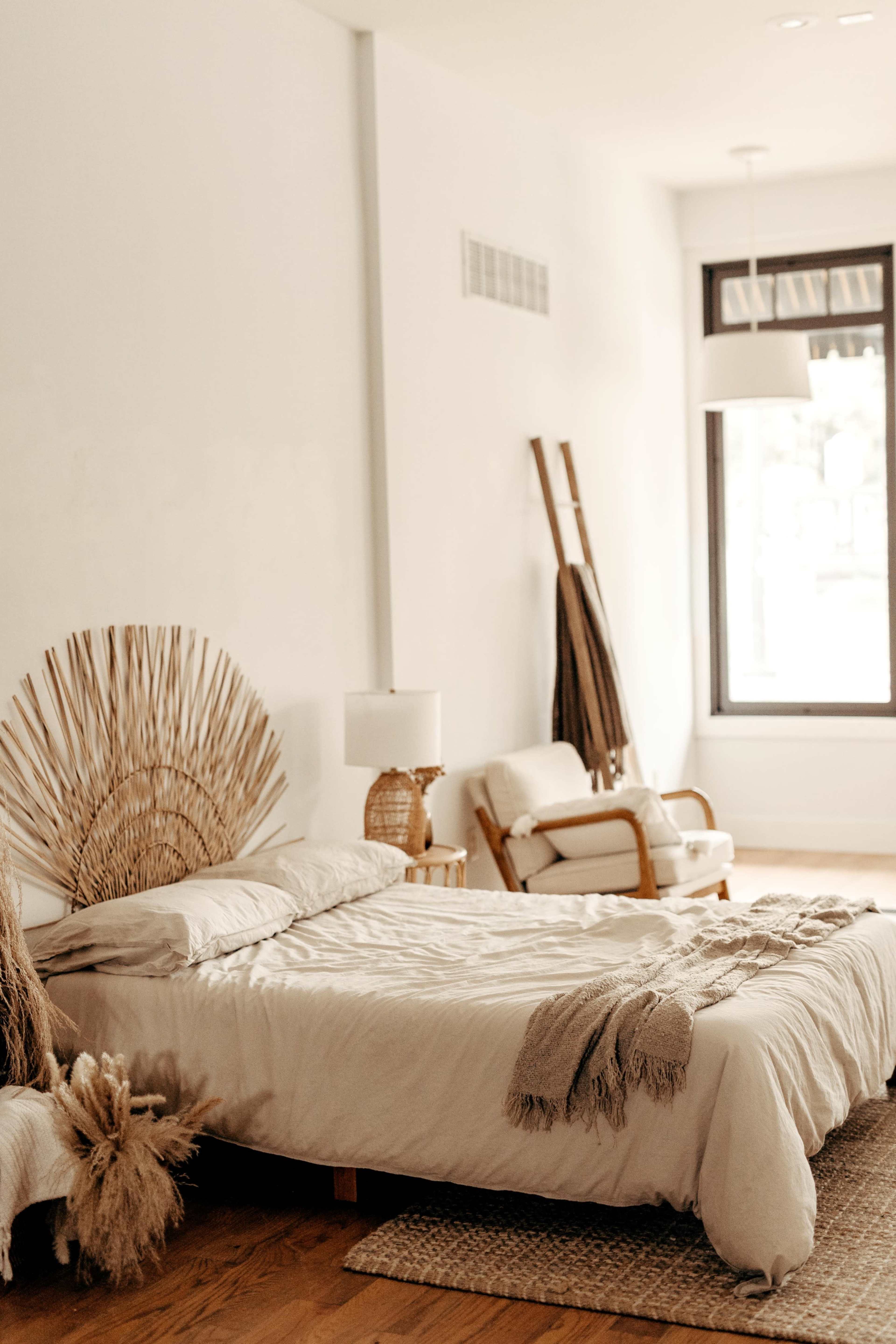The image shows a simple bedroom featuring a bed with a textured headboard, a chair, and natural decor elements, all set in a well-lit space with large windows.