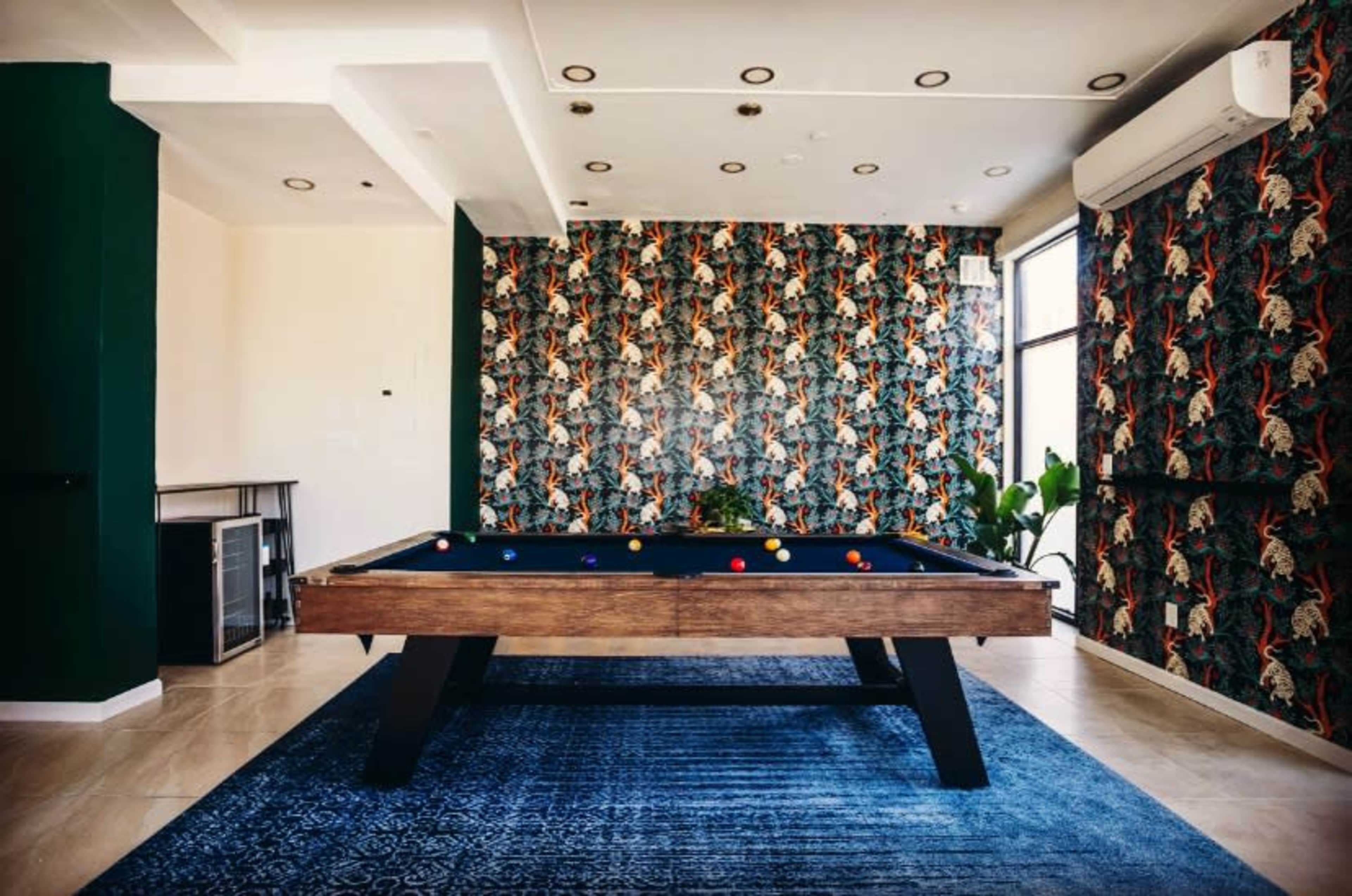 A modern game room featuring a billiards table, patterned wallpaper, and large windows with plants nearby.