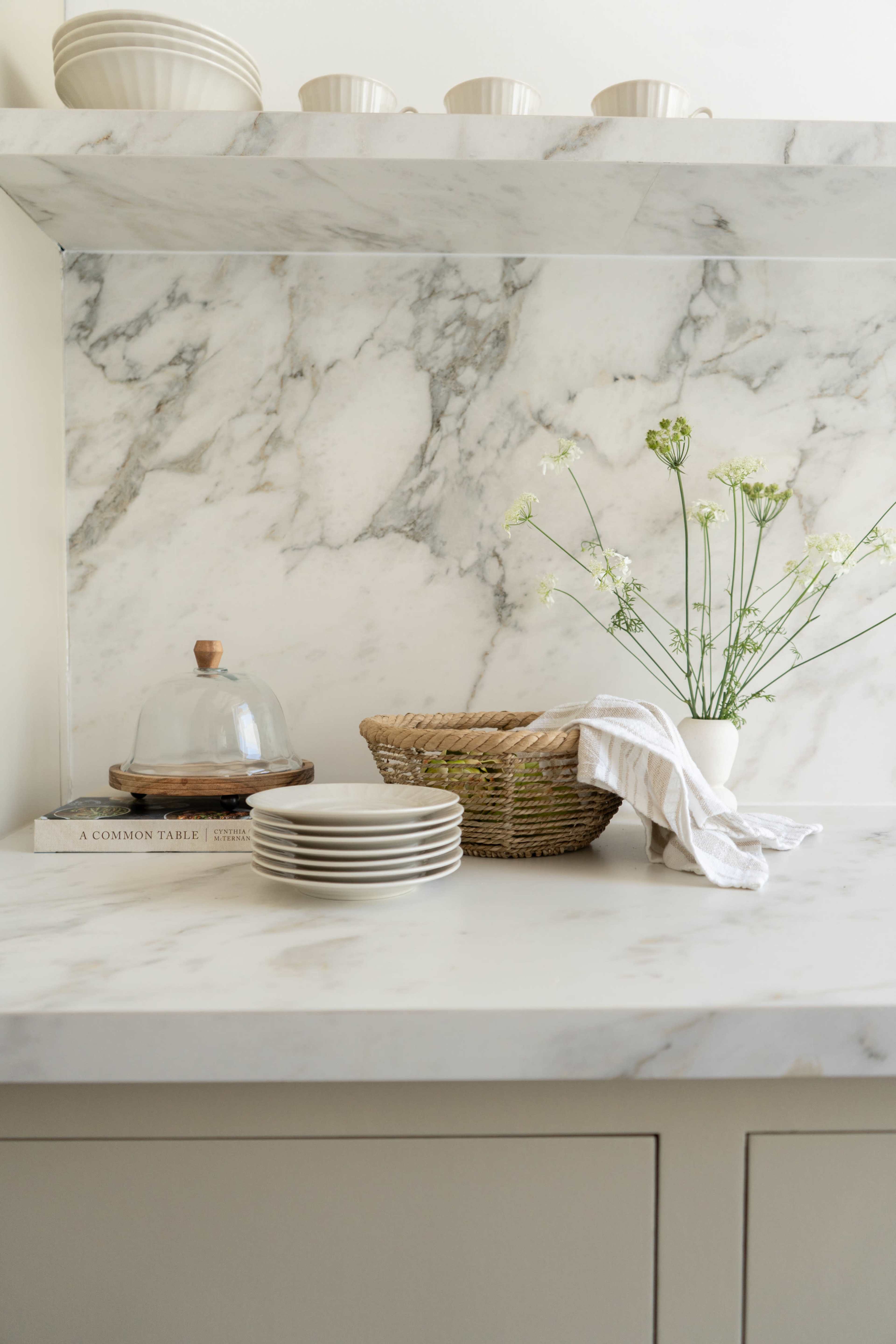 A marble countertop with stacked white plates, a glass cloche, a woven basket, a cloth napkin, and a vase of flowers.