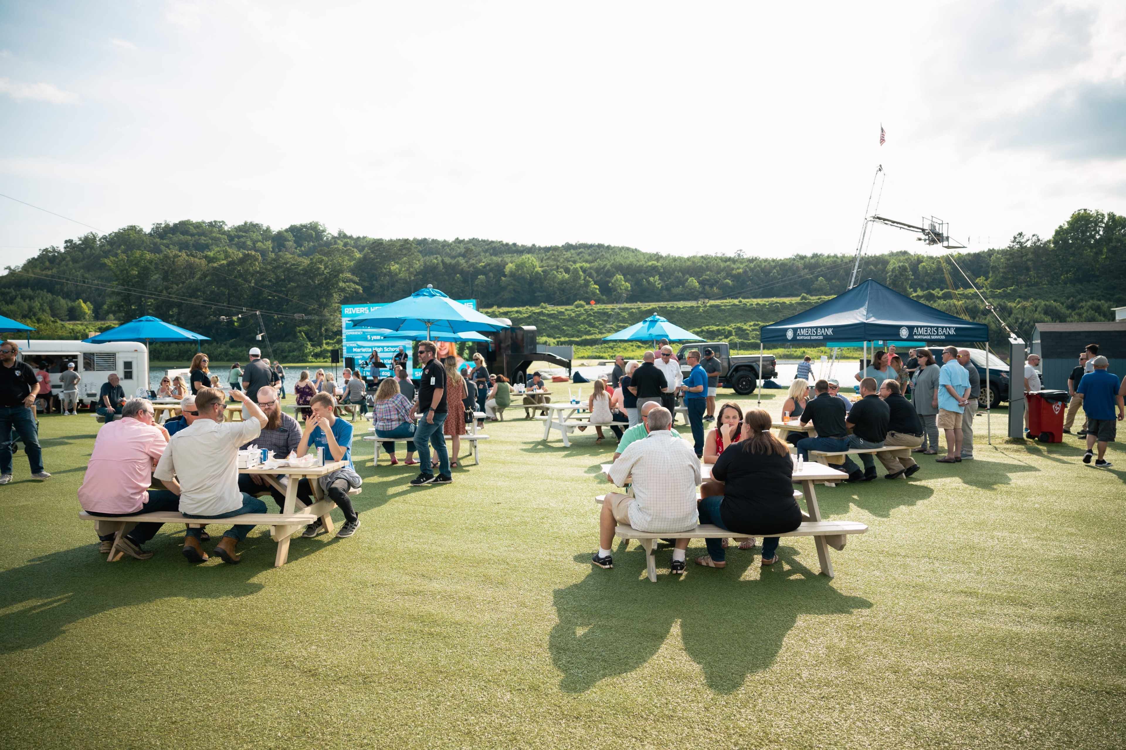 A large group of people socializes around picnic tables at an outdoor event on a green field, with tents and a scenic background.