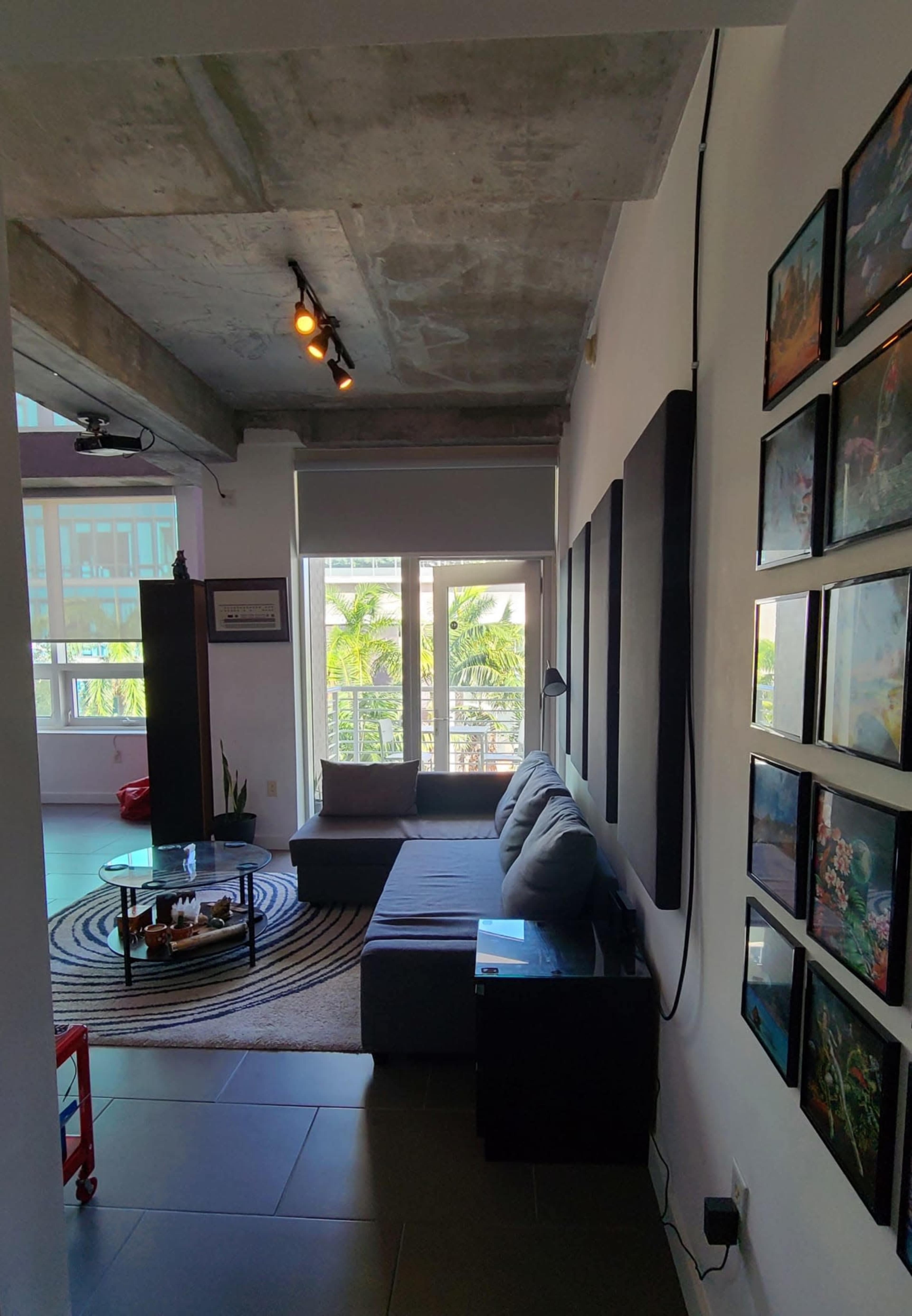 A living room features a gray sofa, a round coffee table on a striped rug, and a wall of framed pictures beside large windows with views of greenery.