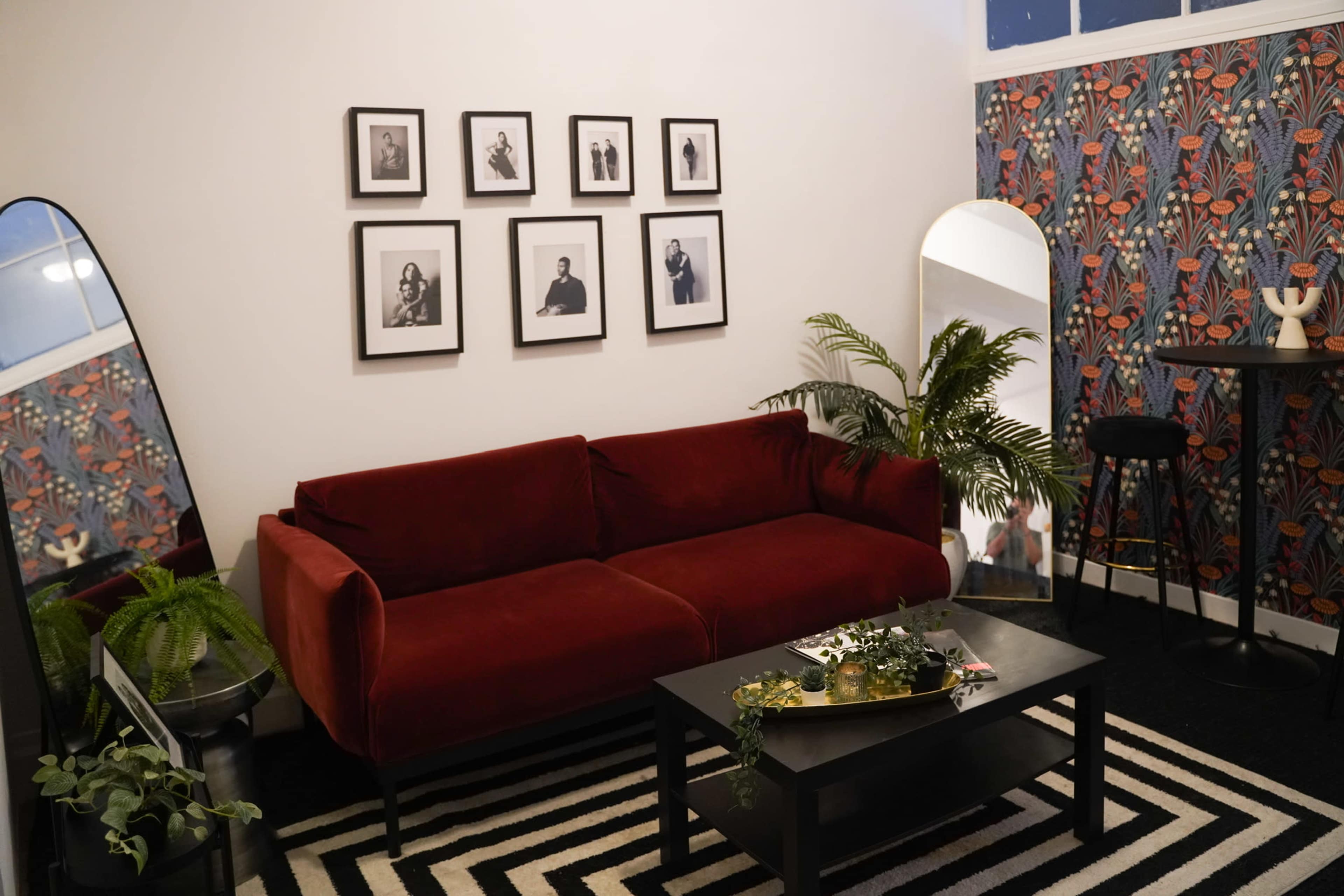A modern living room with a red sofa, a black coffee table, framed photographs on the wall, a large mirror, and decorative plants.