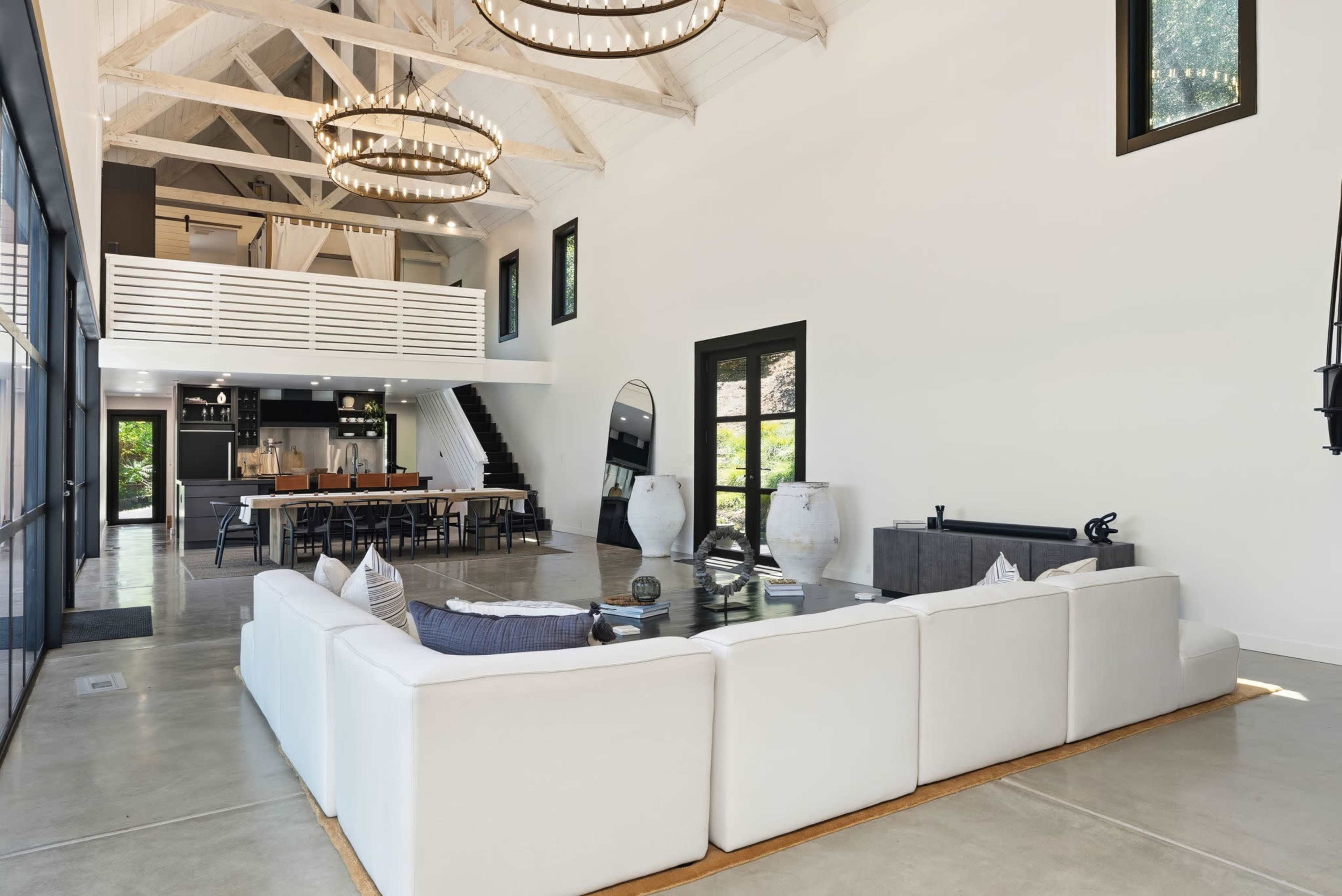 The image shows a spacious, modern living area featuring a white sectional sofa, high ceilings with wooden beams, large windows, and an adjacent dining area with a long table.
