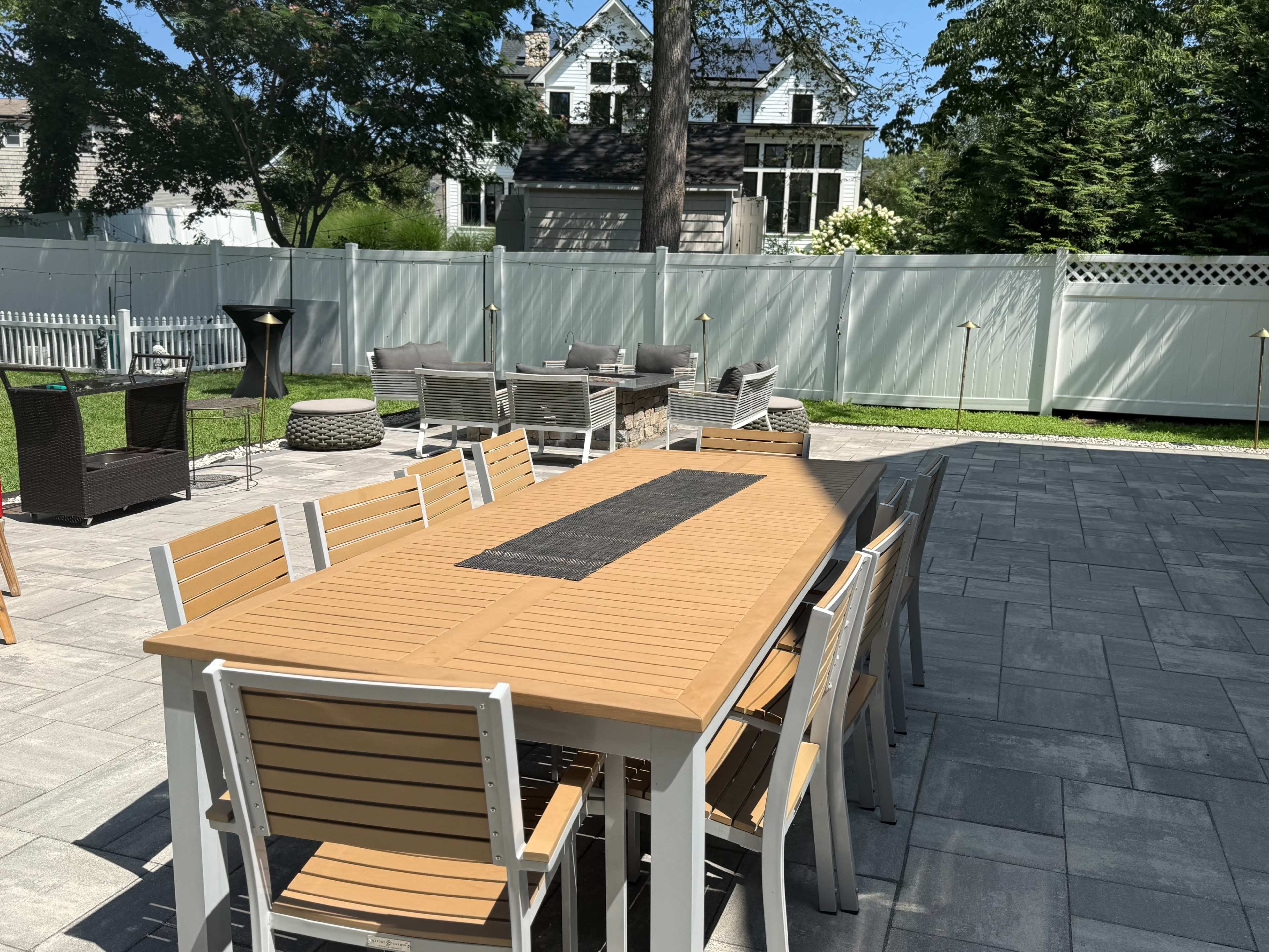 A large outdoor dining table with multiple chairs is set on a stone patio, surrounded by a well-maintained yard and a backdrop of houses and trees.