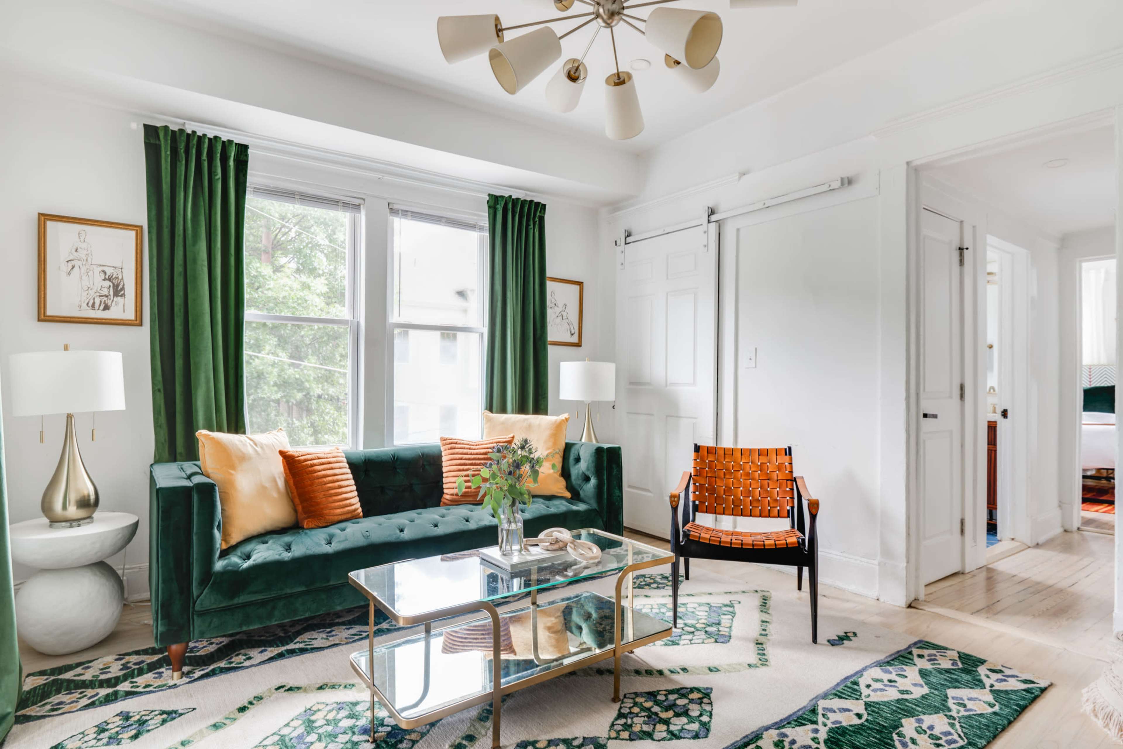 The scene features a well-lit living room with a green velvet sofa, an orange accent chair, a glass coffee table, and two white lamps beside the window, all set against a patterned rug.