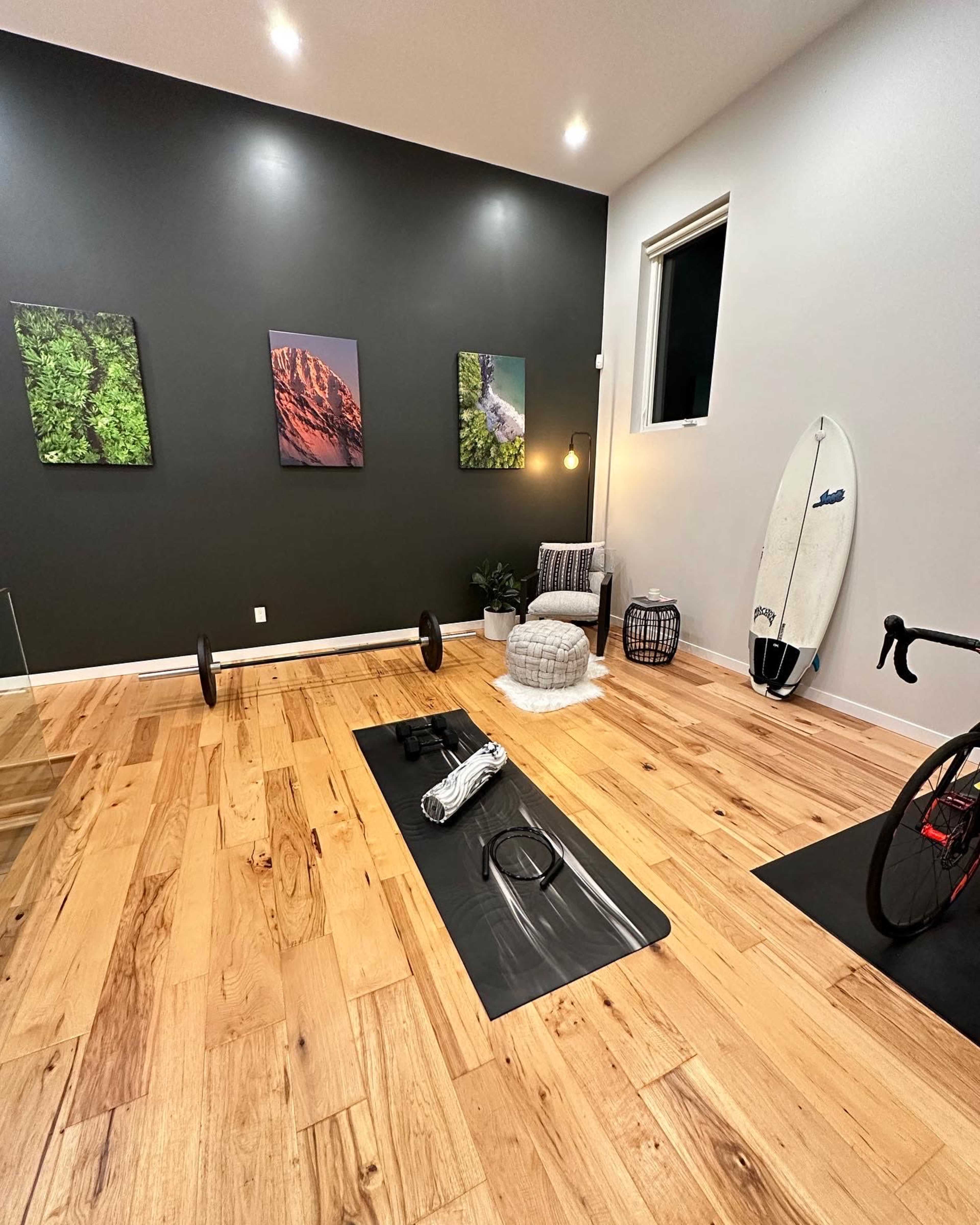 The room features wooden flooring with a weight bar, workout mat, bicycle, surfboard, and artwork on the walls.