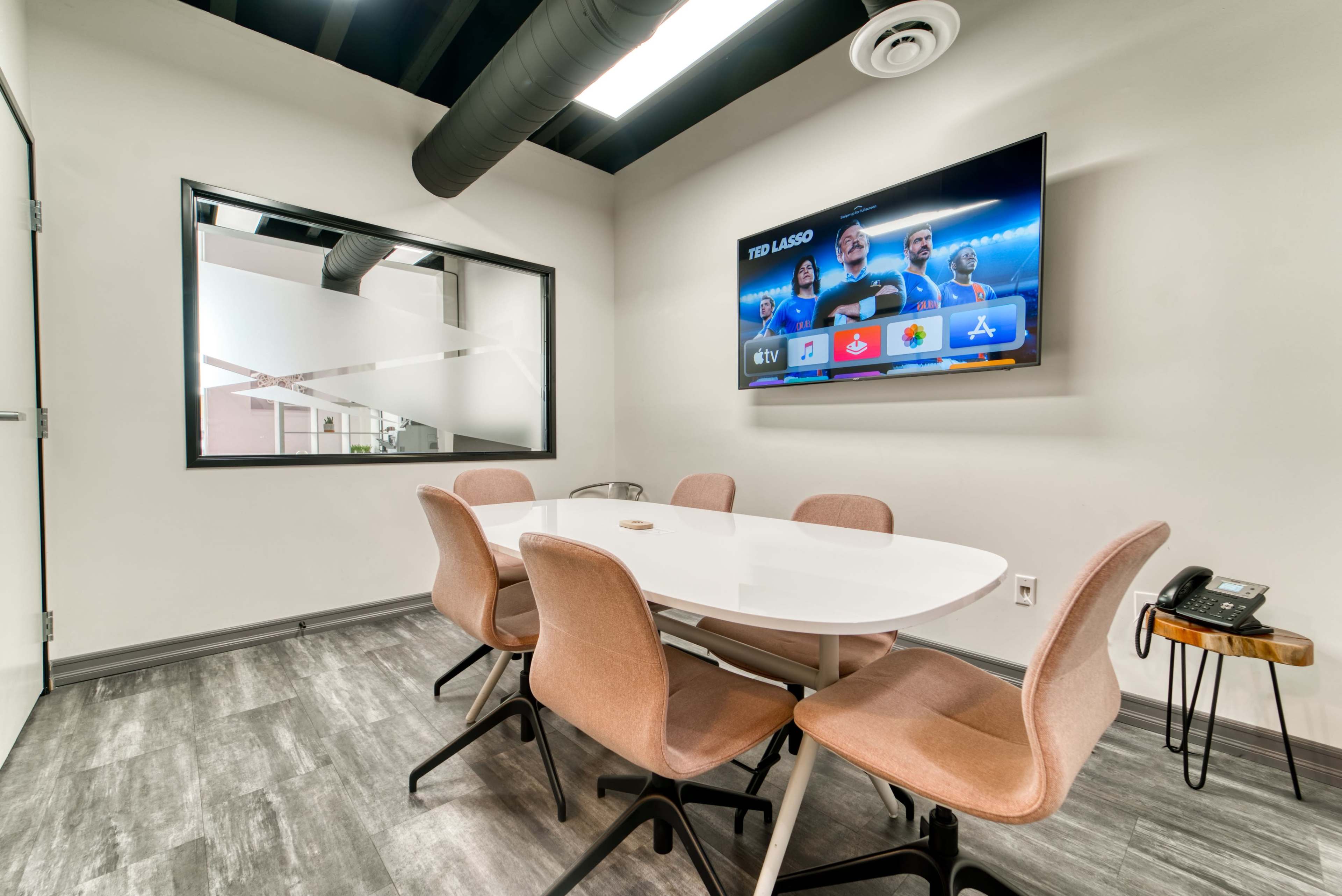 A modern conference room with a round white table, six beige chairs, a wall-mounted TV displaying a screen, and a landline phone on a side table.