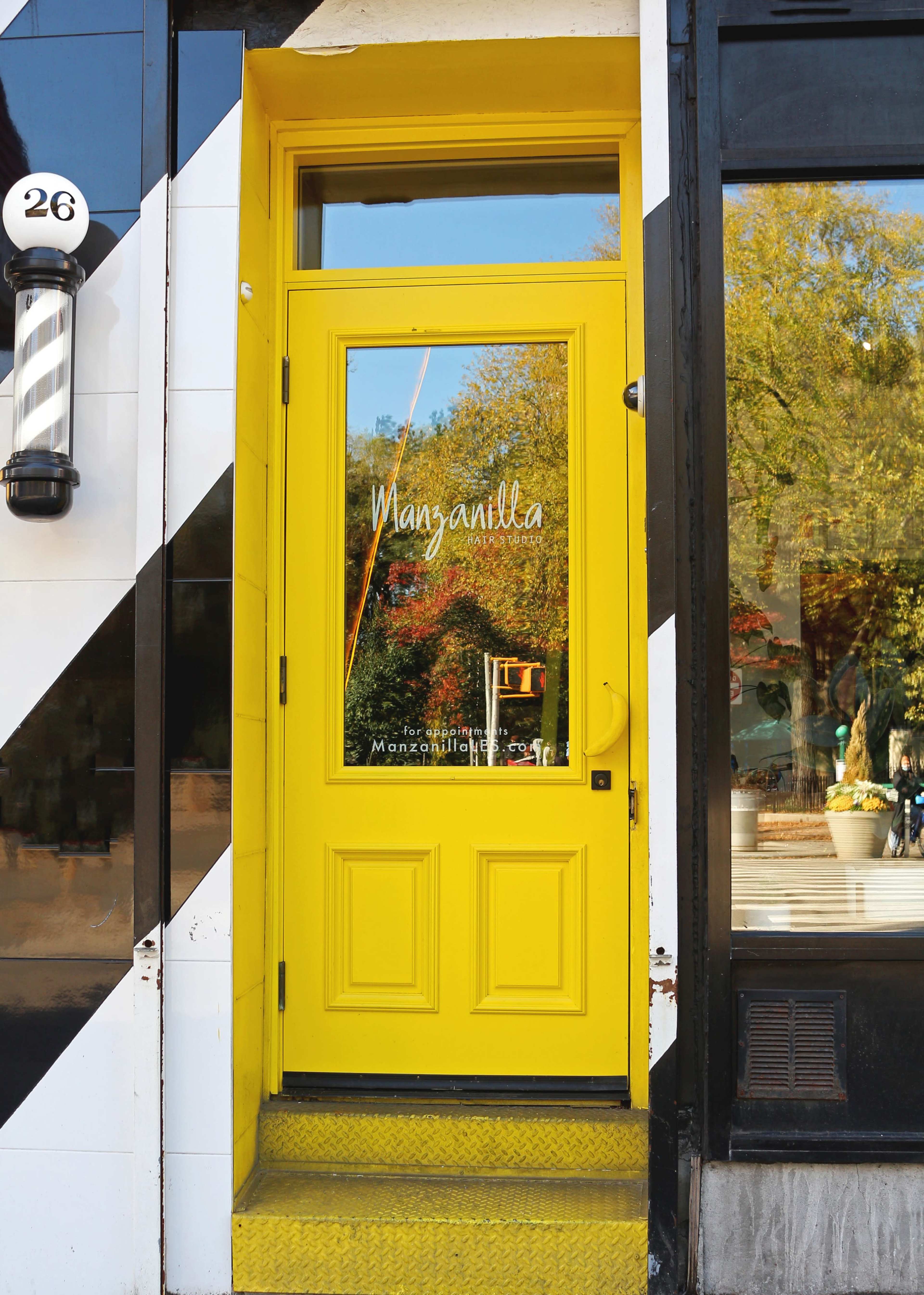 The image shows a vibrant yellow door with the name "Manzanilla" on it, framed by a black and white striped exterior.