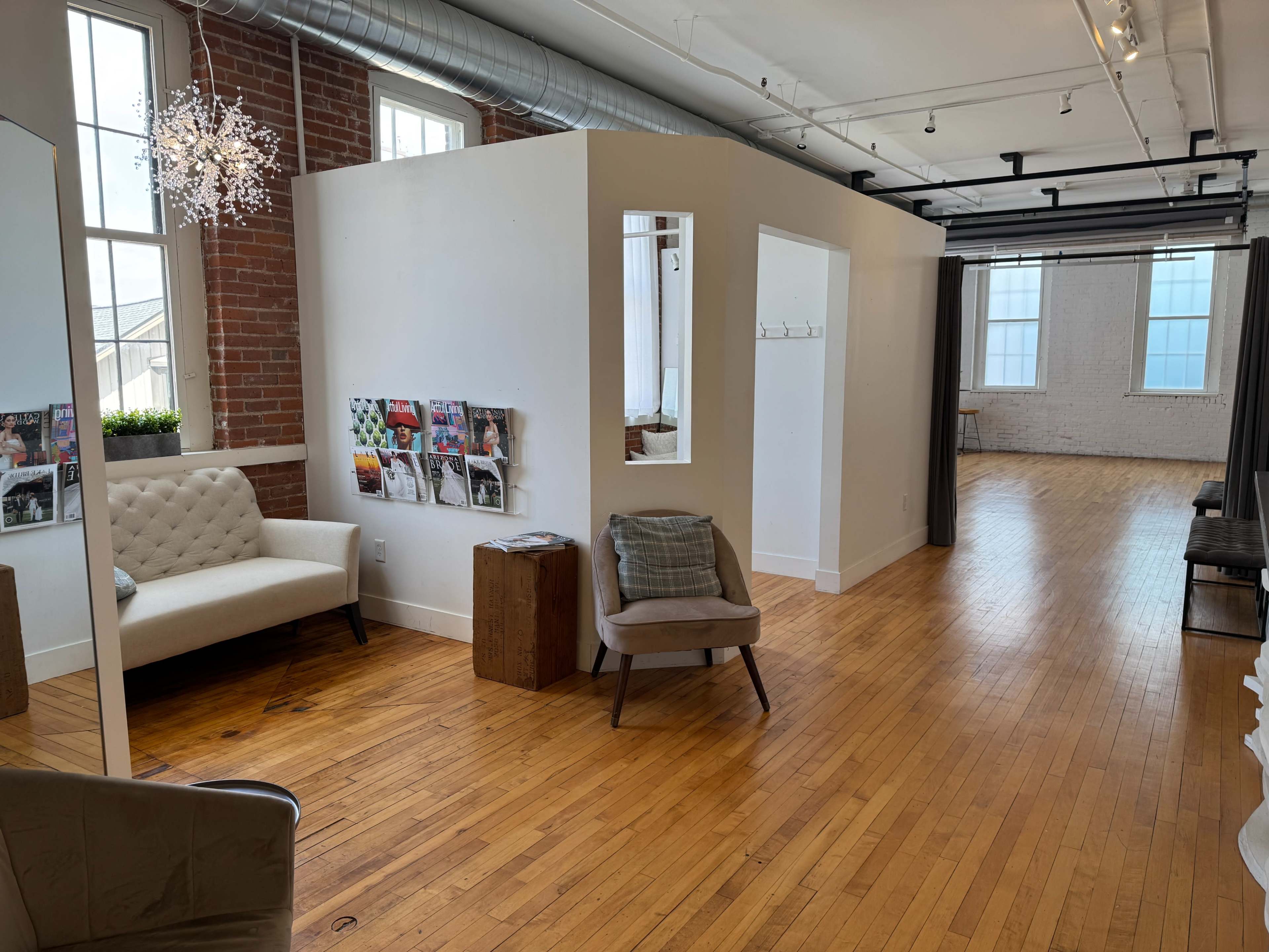 The image shows a bright, modern studio space with wooden floors, white walls, and a cozy seating area featuring a tufted beige sofa and a chair, along with a magazine rack on the wall.