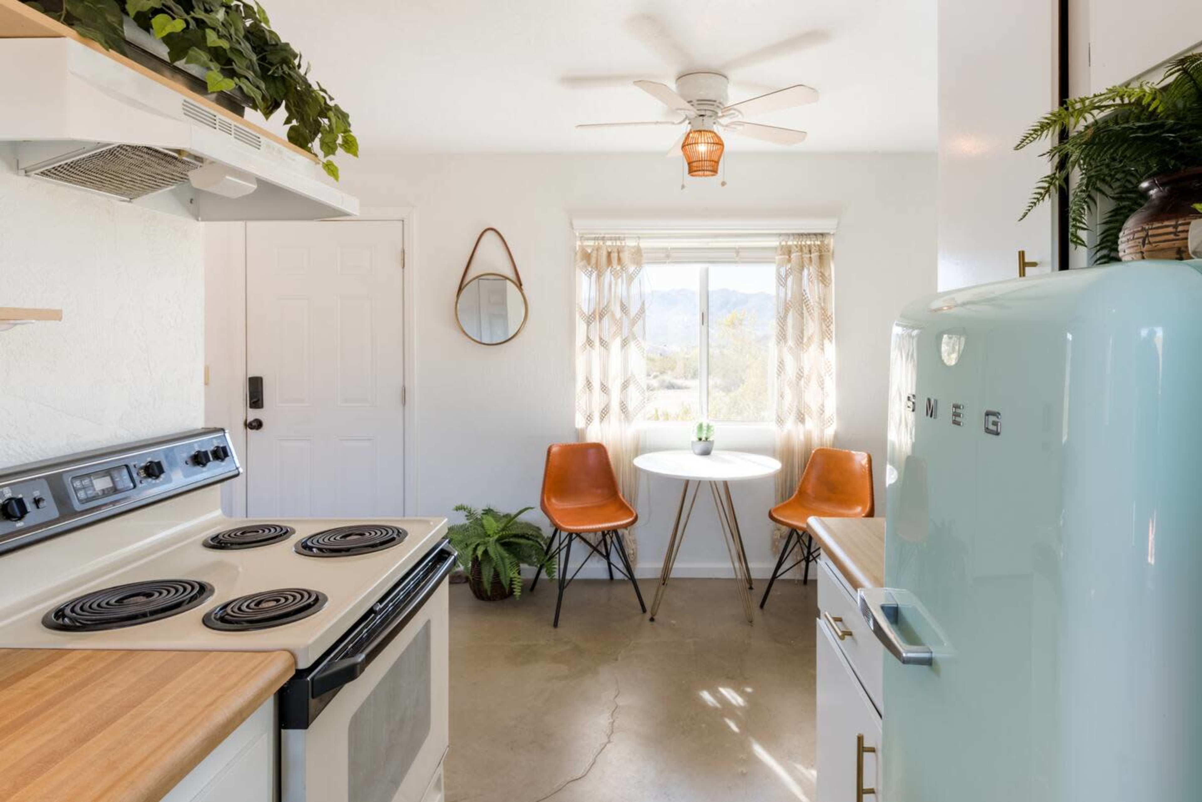 A bright kitchen features a stove, a retro refrigerator, a round table with two chairs, and a window with mountain views in the background.
