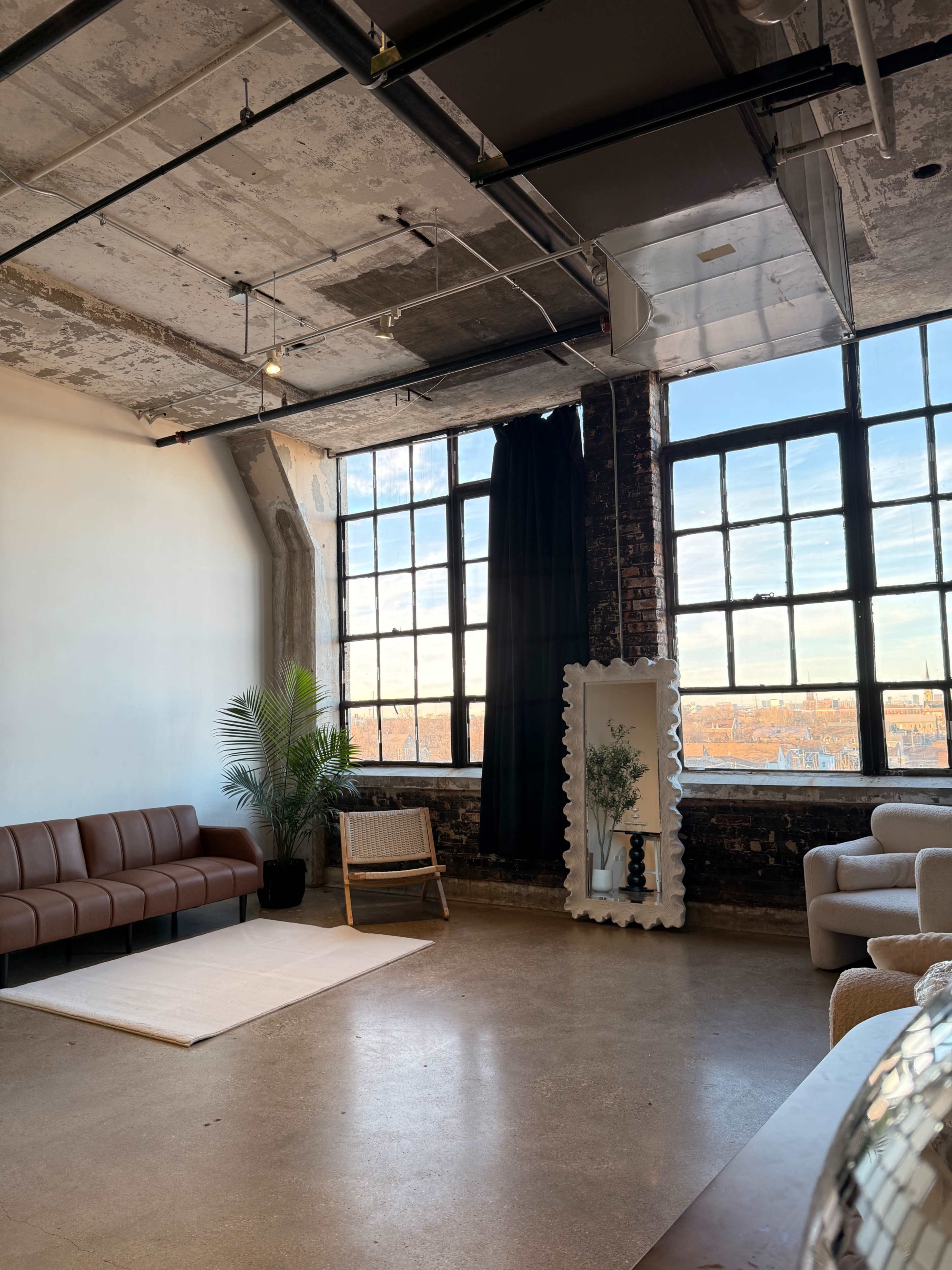 The image shows a spacious room featuring a brown leather sofa, a light rug on the floor, large windows with a city view, and a white decorative mirror with a plant nearby.