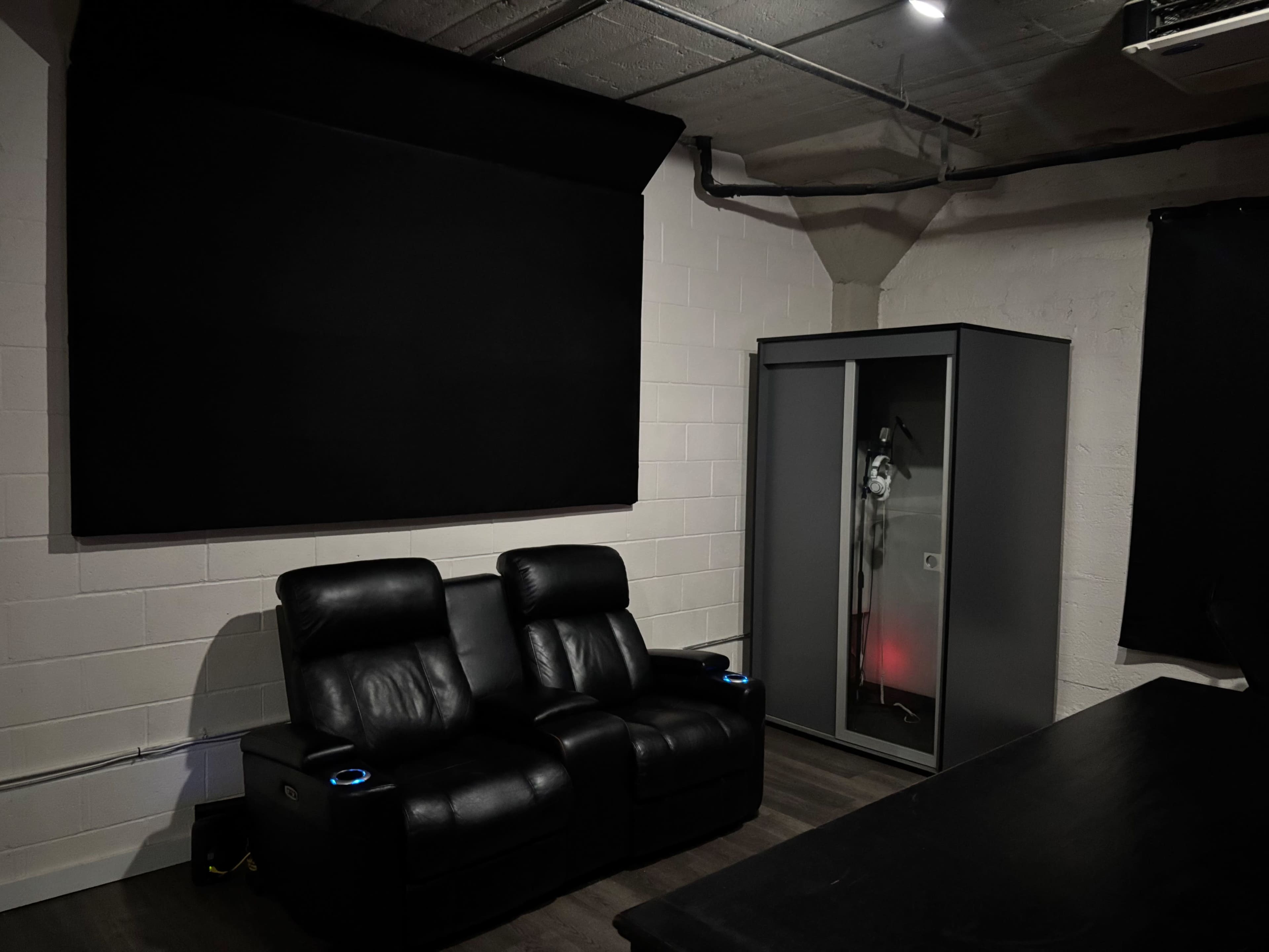 The image shows a small room with two black reclining chairs facing a large dark screen, and a glass display cabinet containing a figure, illuminated with red light, against a white brick wall.