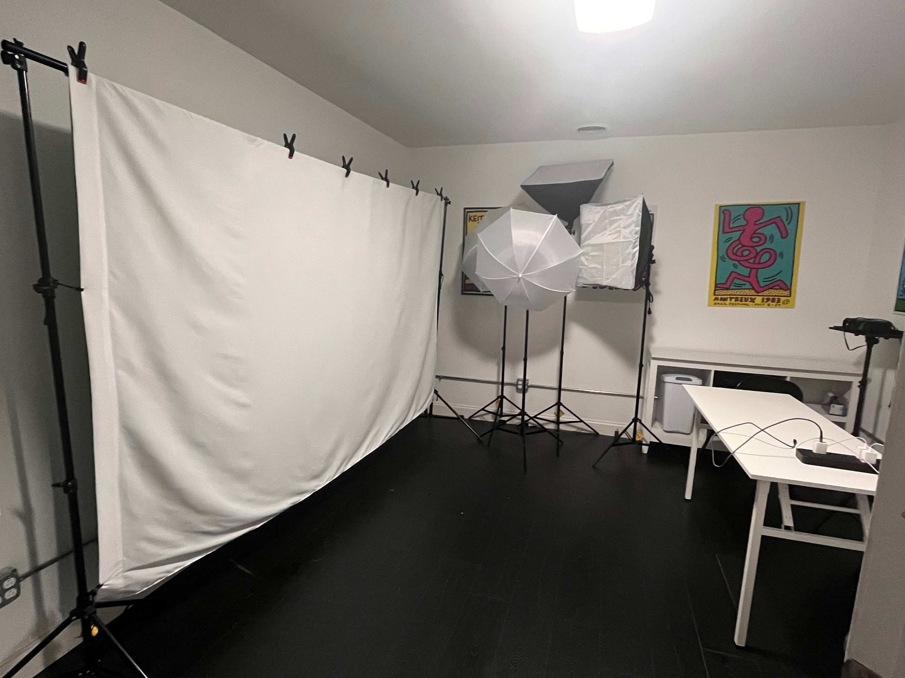 The image shows a photography studio setup featuring a white backdrop, lighting equipment, and a table against a plain wall.