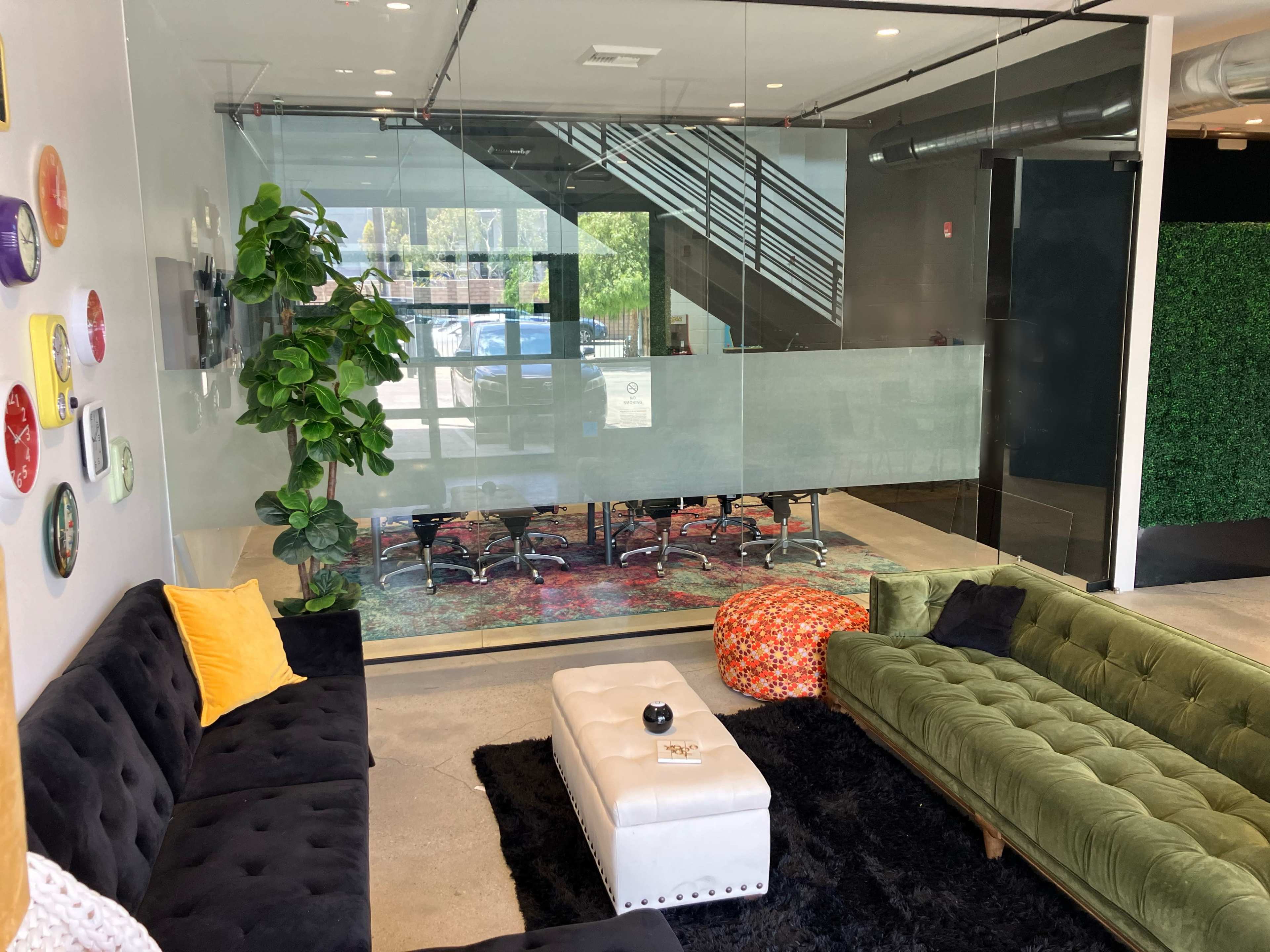 A modern office space features a glass-walled conference room with a large table surrounded by chairs, adjacent to a cozy lounge area with sofas and decorative plants.