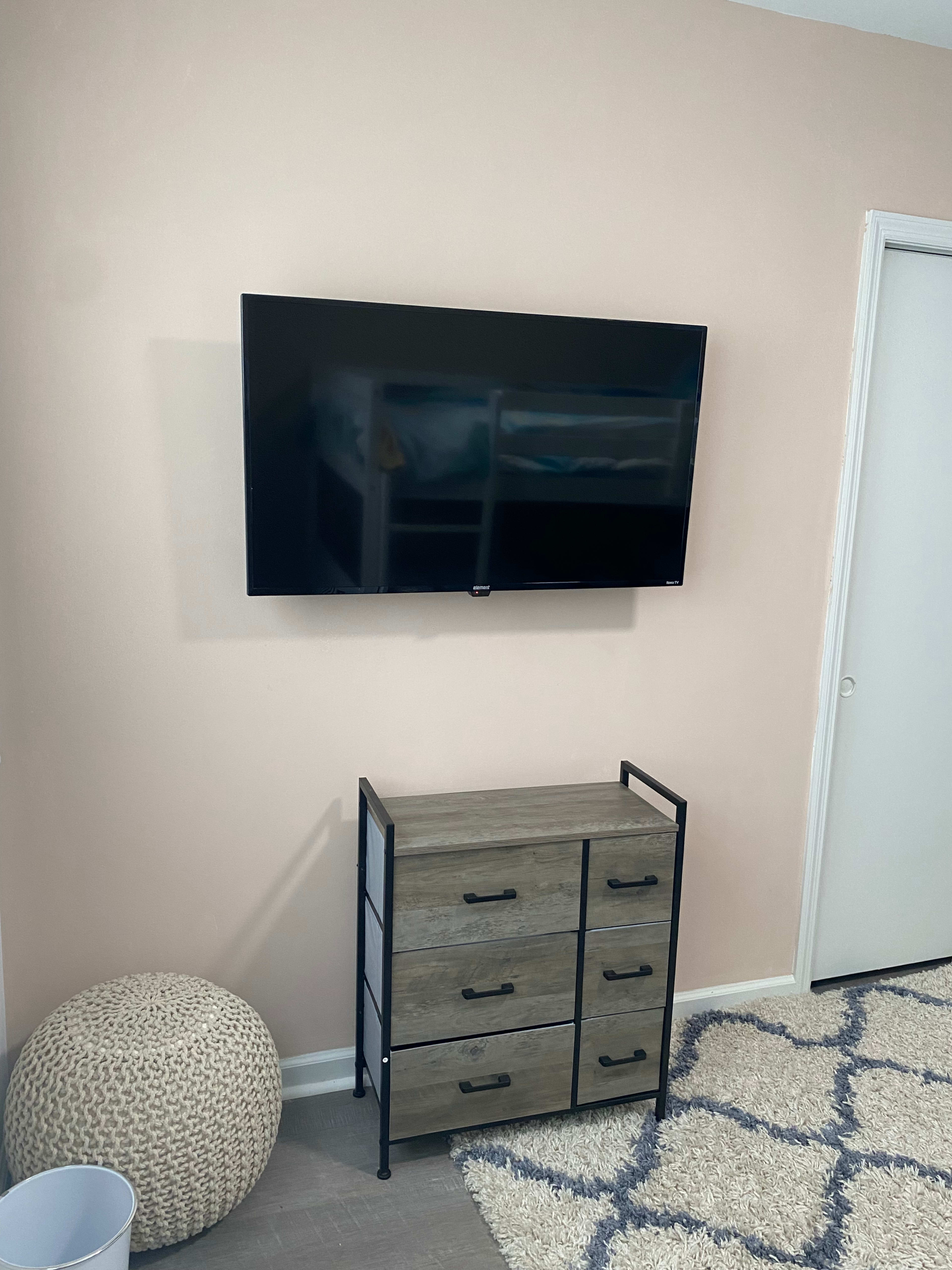 A wall-mounted television is positioned above a wooden dresser with four drawers, adjacent to a white door and a round pouf on the floor.