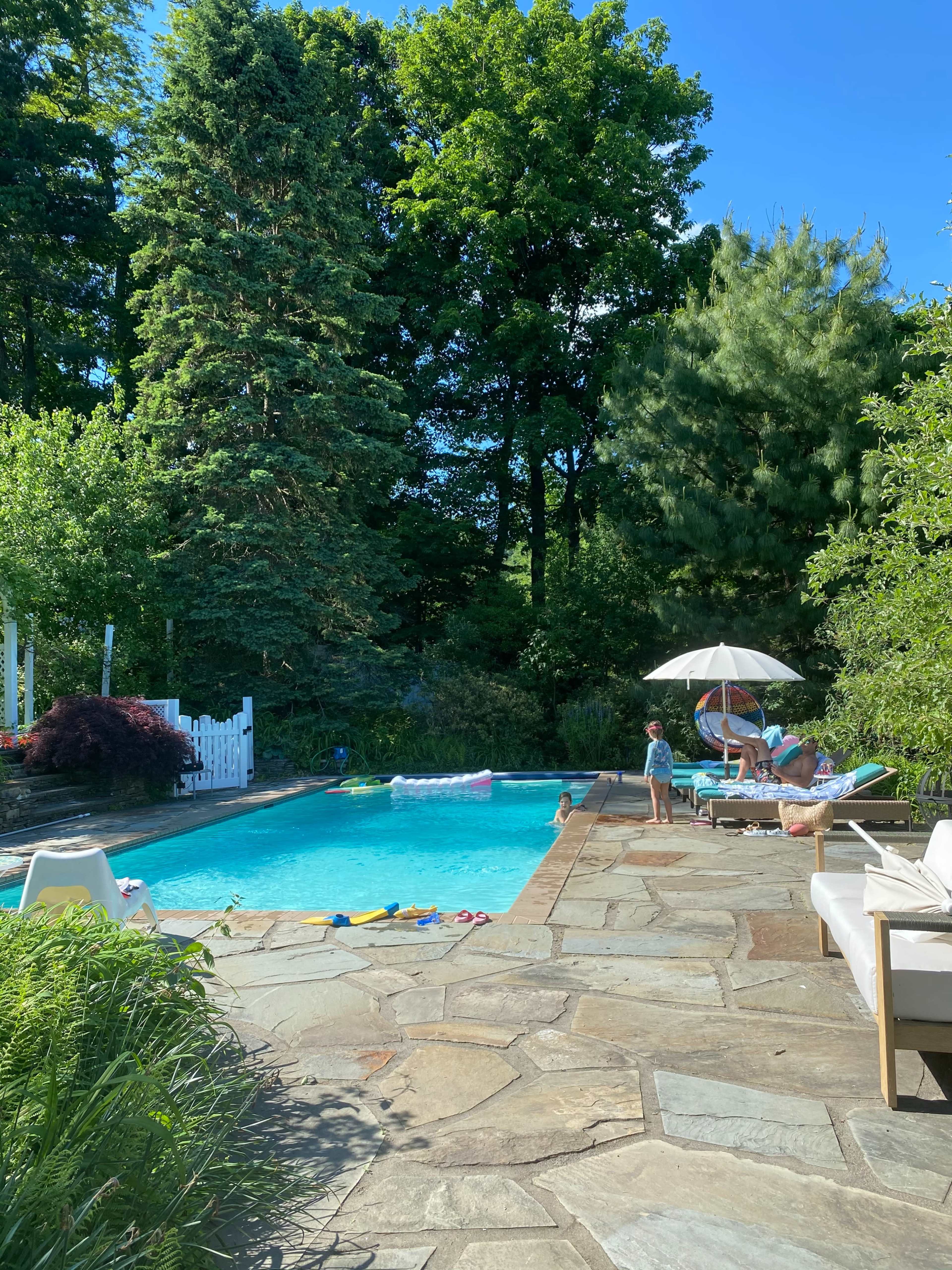 A family enjoys a sunny afternoon by a swimming pool surrounded by lush trees and stone patio.