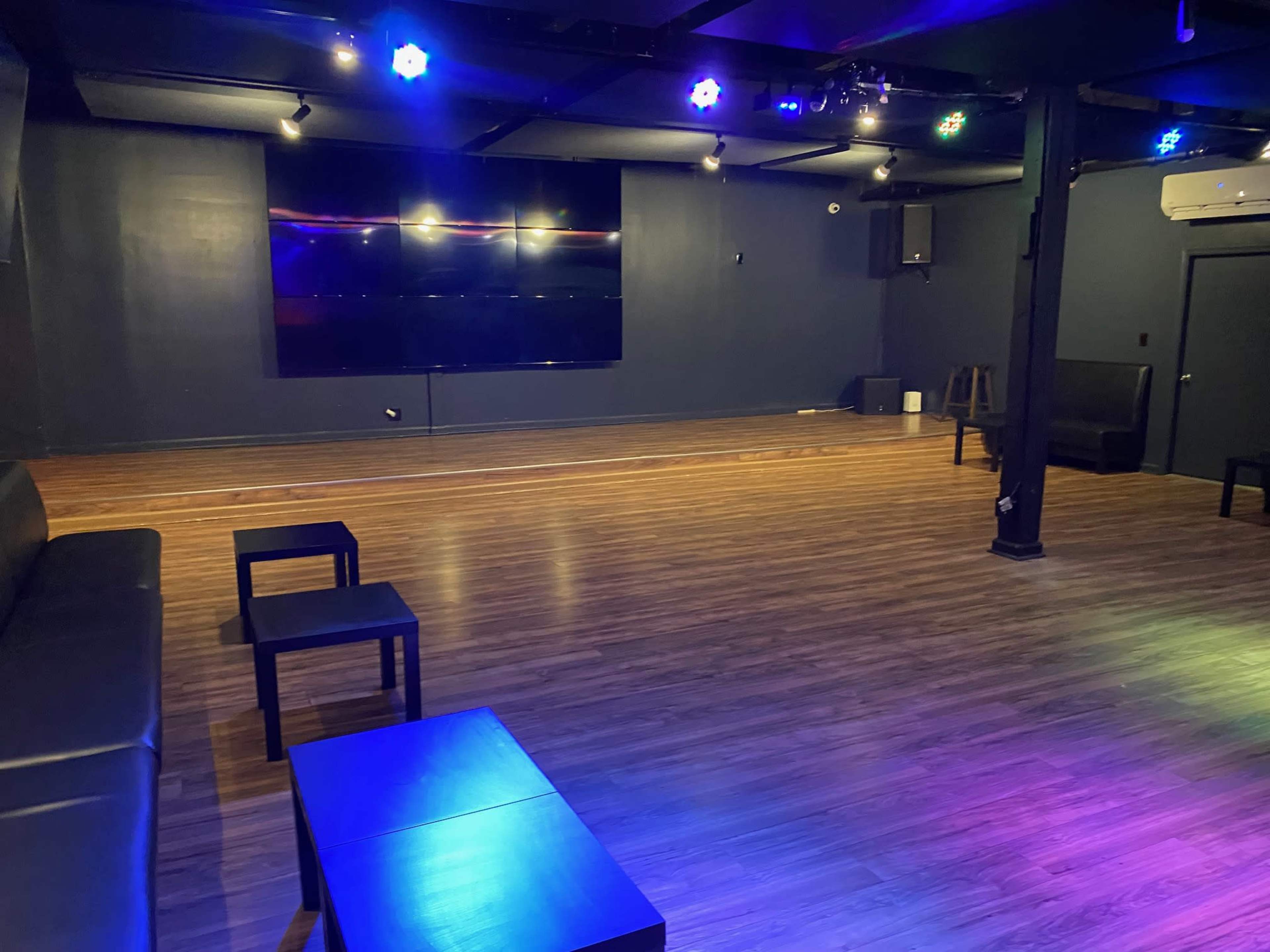 A spacious, empty room with a wooden floor, black seating, and a large screen on the wall, illuminated by multicolored lights.