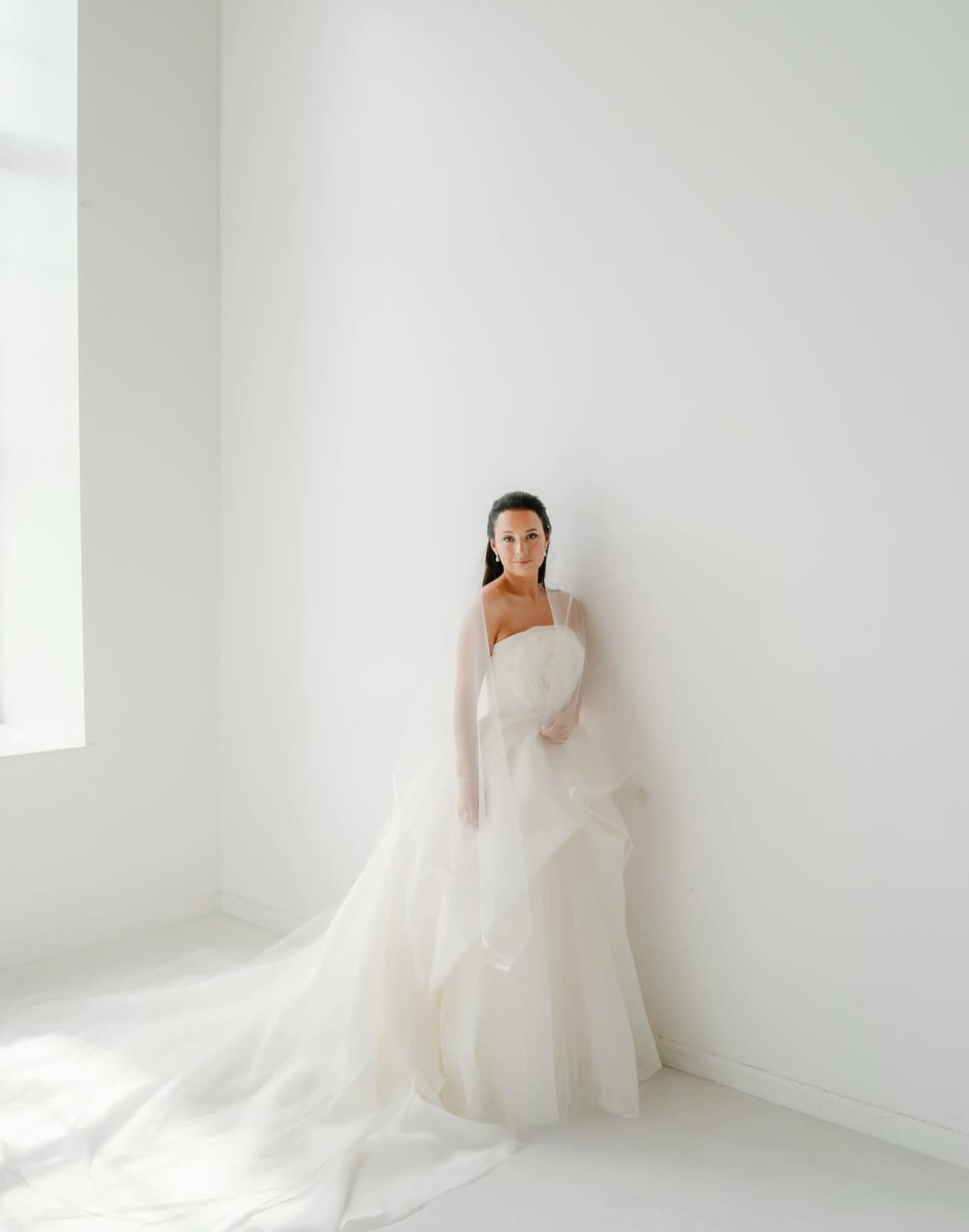 A woman in a flowing white gown stands against a plain white wall.