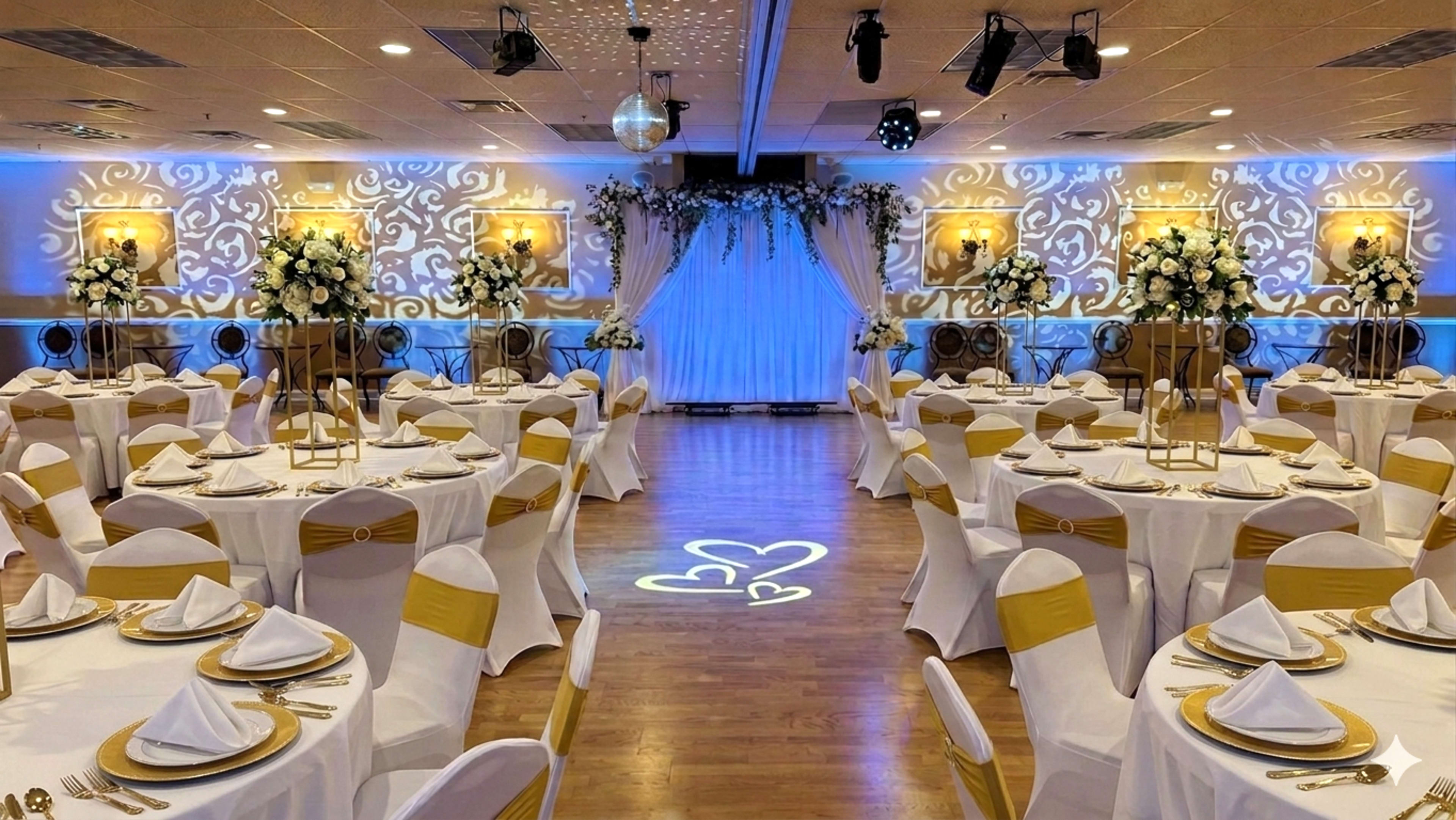 The image shows a lavish banquet hall set up for an event, featuring circular tables covered with white tablecloths and gold accents, floral decorations, and a backdrop with white drapery.