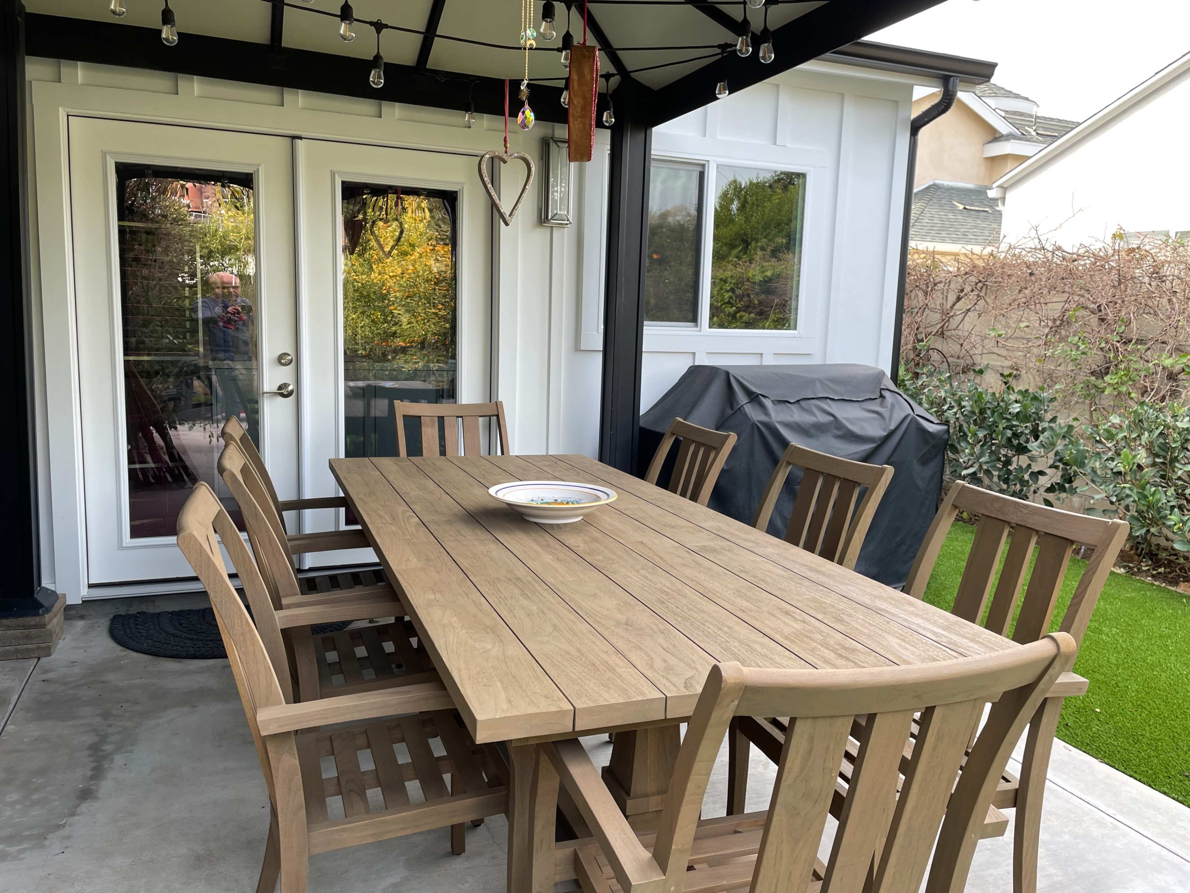 A wooden dining table with eight chairs is set under a covered patio, accompanied by a grill and surrounded by greenery.