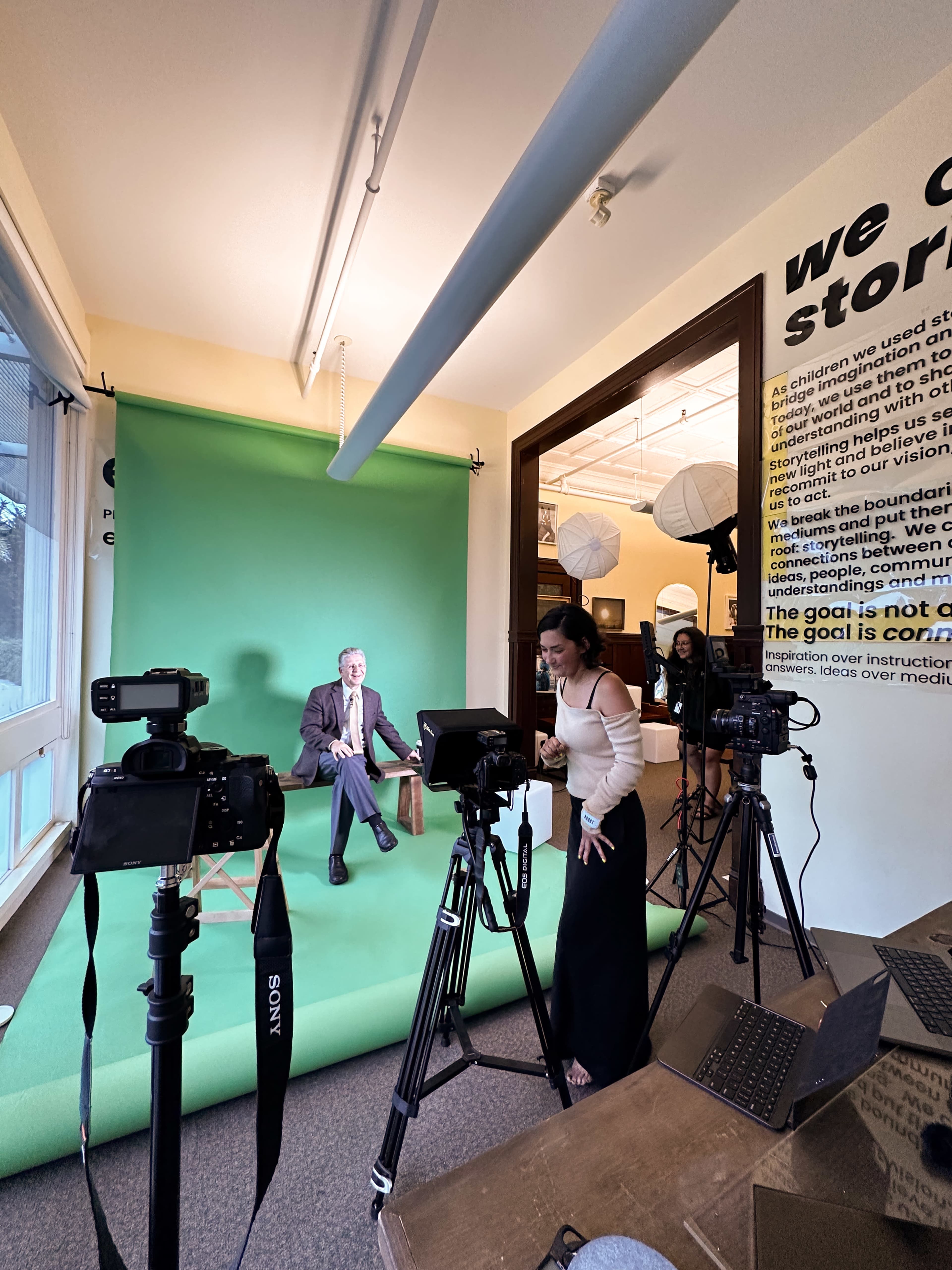 A man sitting on a stool in front of a green screen is being filmed by a woman with multiple cameras set up around them.