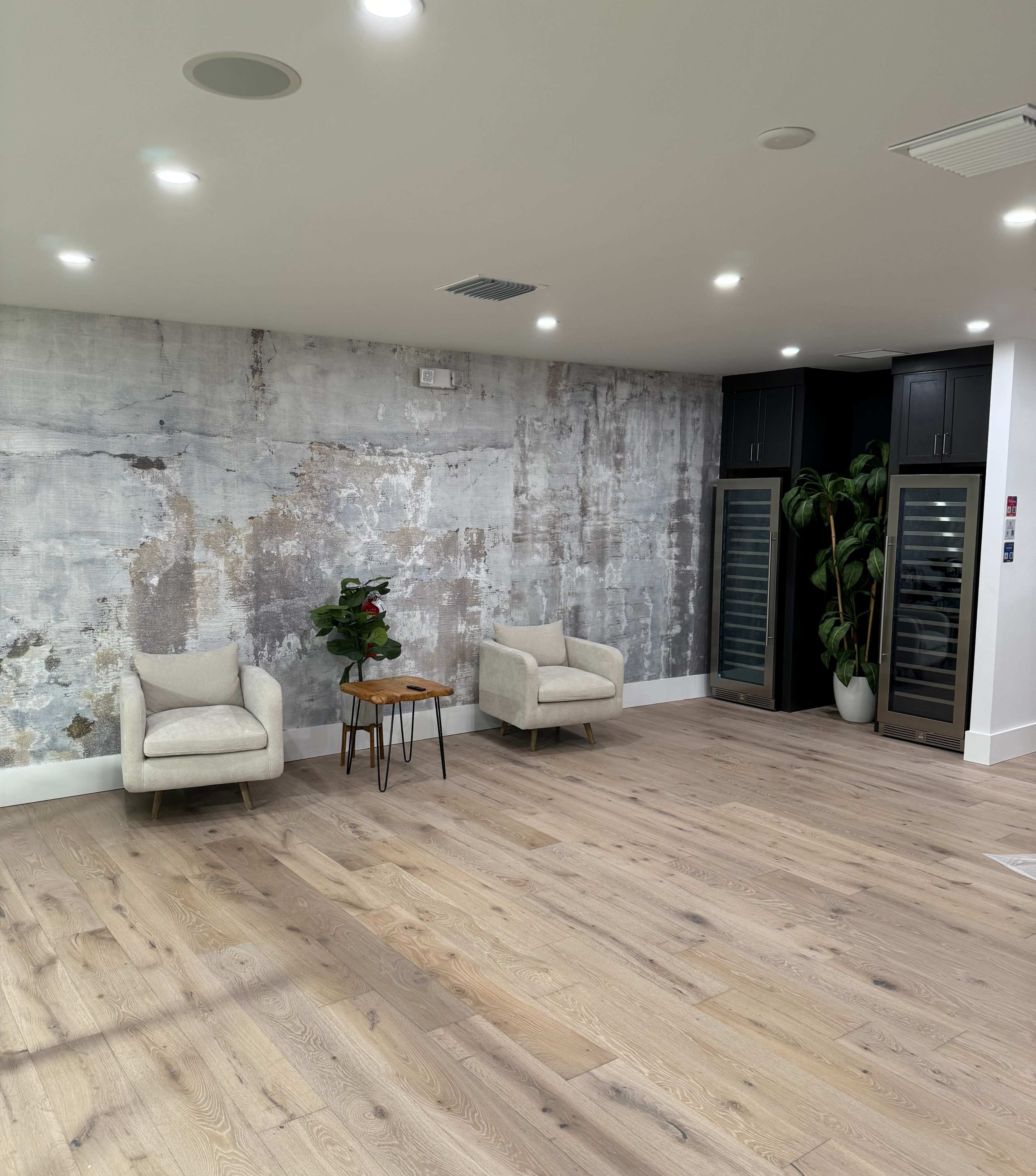 A spacious room with two light-colored armchairs, a small wooden table, and a textured gray wall, along with two glass-fronted storage units and a tall plant in the corner.
