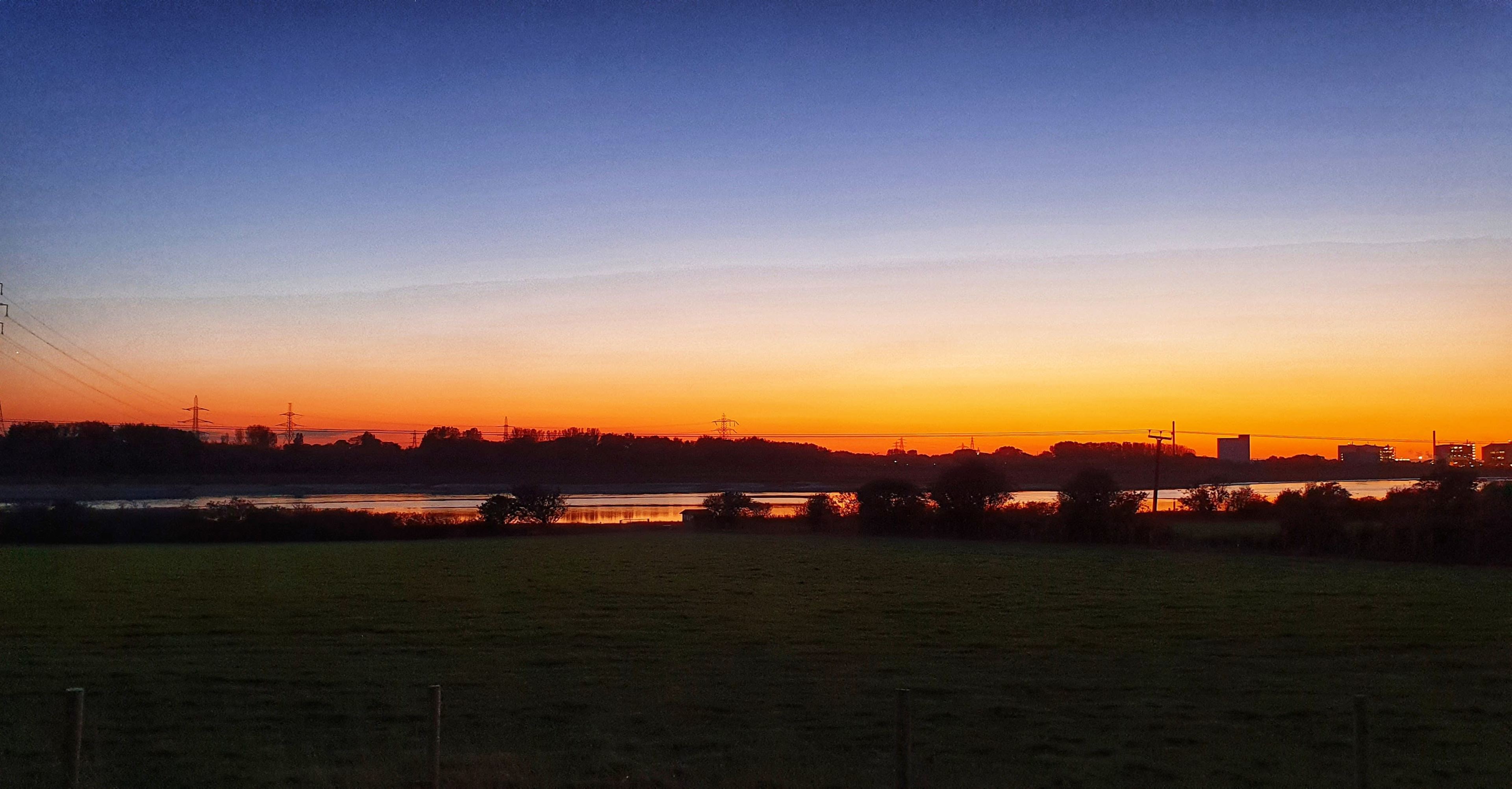 A colorful sunset over a body of water, with silhouettes of trees and power lines in the foreground.
