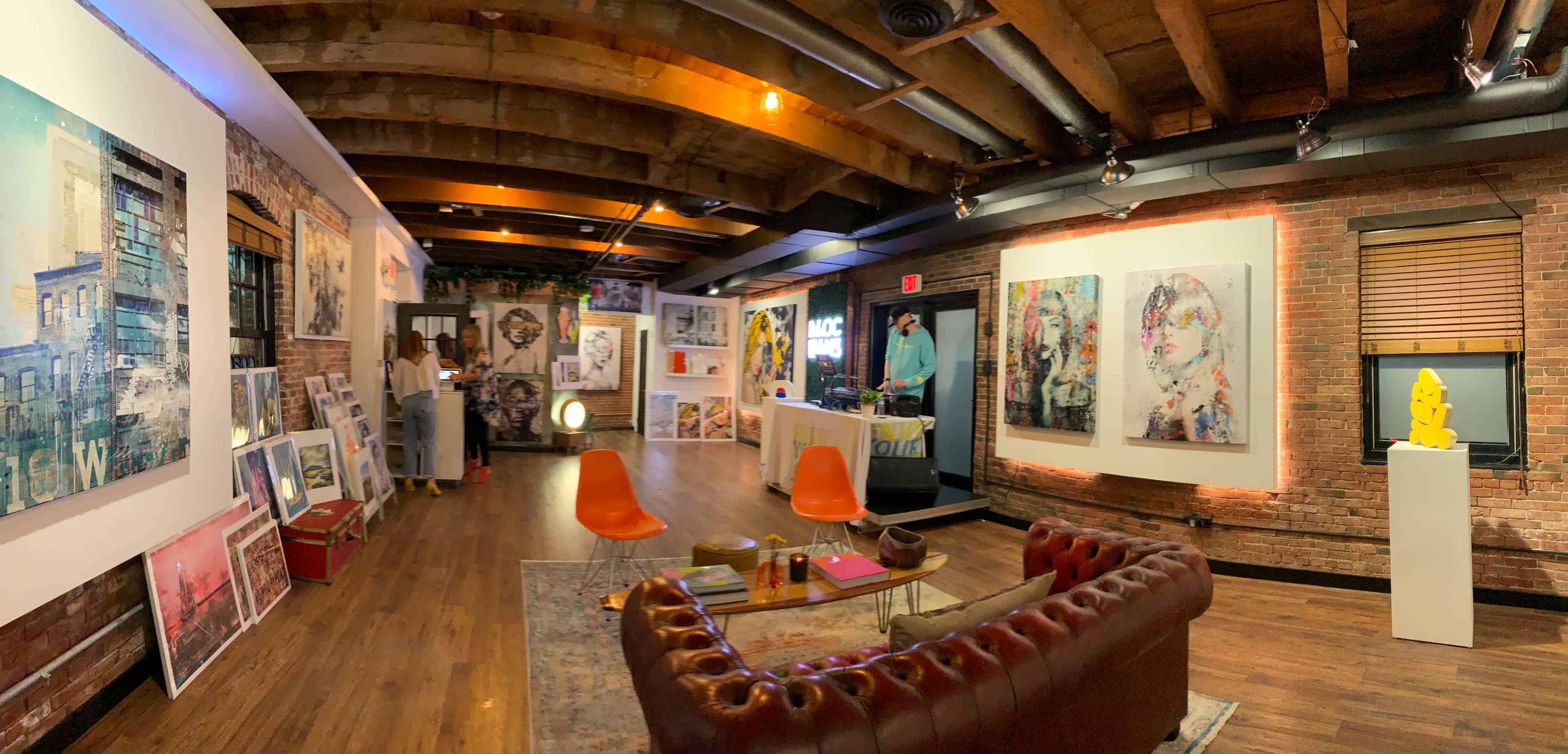 An art gallery with exposed brick walls and wooden beams, featuring various artworks displayed on the walls and a seating area with orange chairs and a leather couch.