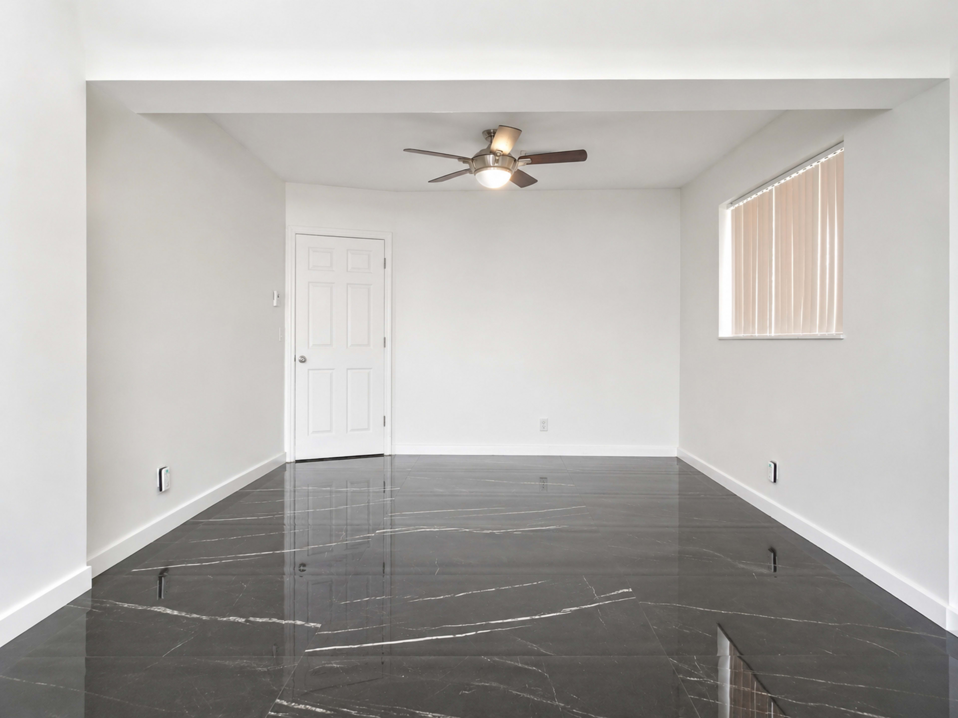 The image shows a spacious room with black polished floors, a ceiling fan, a door, and a window with blinds.