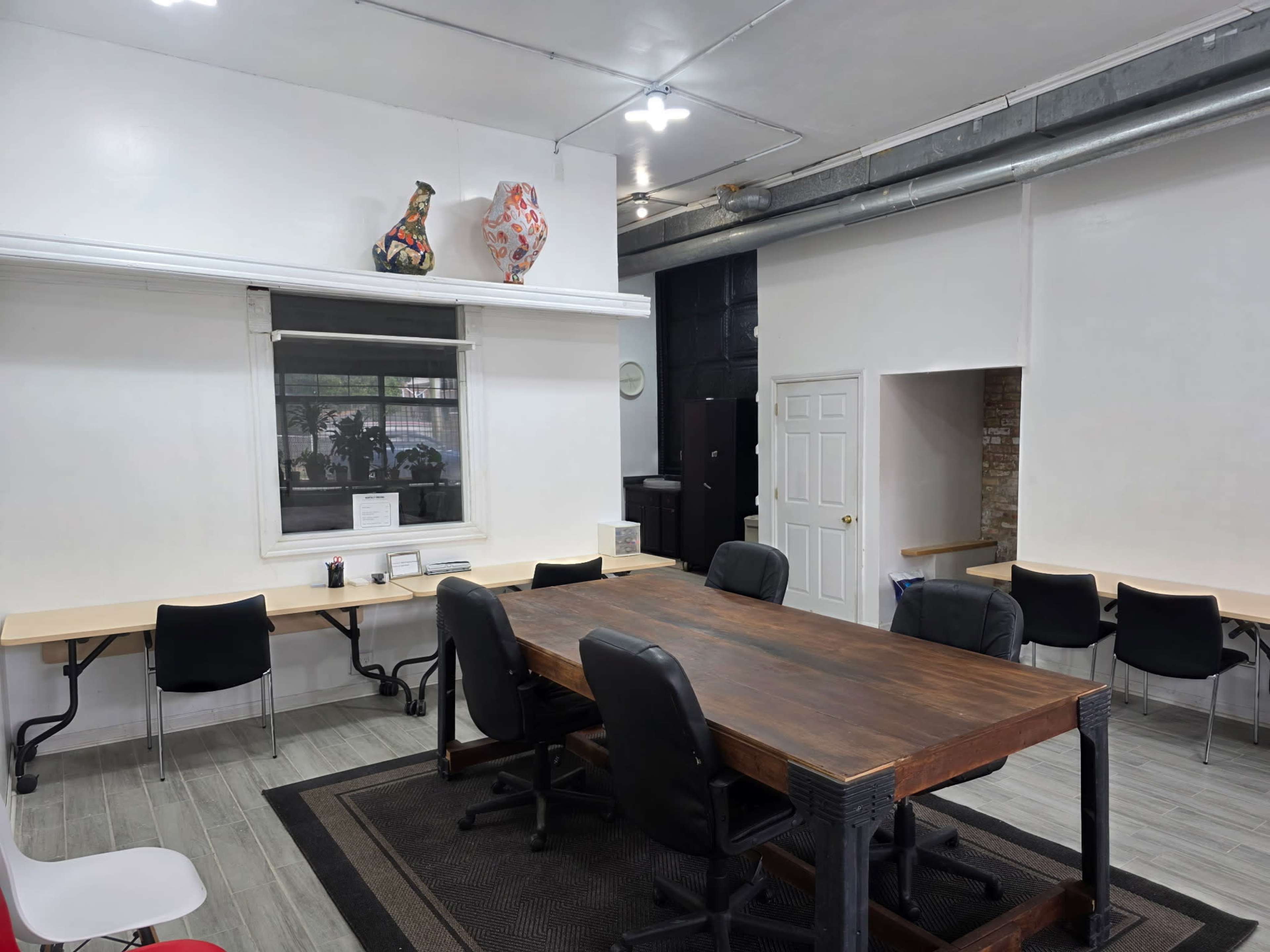 The image shows an office space with a large wooden table in the center, surrounded by black chairs, and additional workstations along the walls.