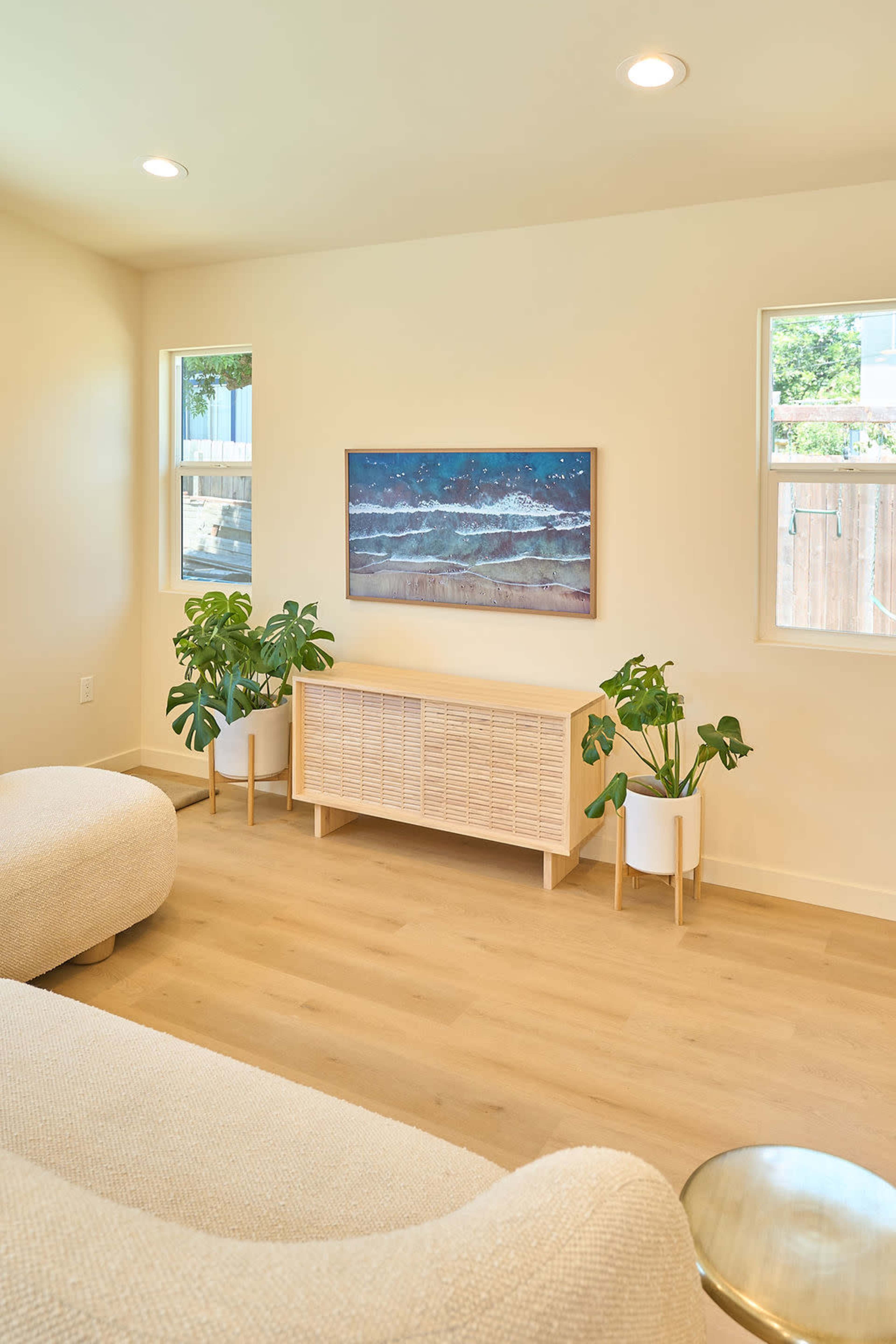 The image shows a light-colored living room featuring a low wooden TV console beneath a beach-themed painting, with two potted plants and soft furniture.