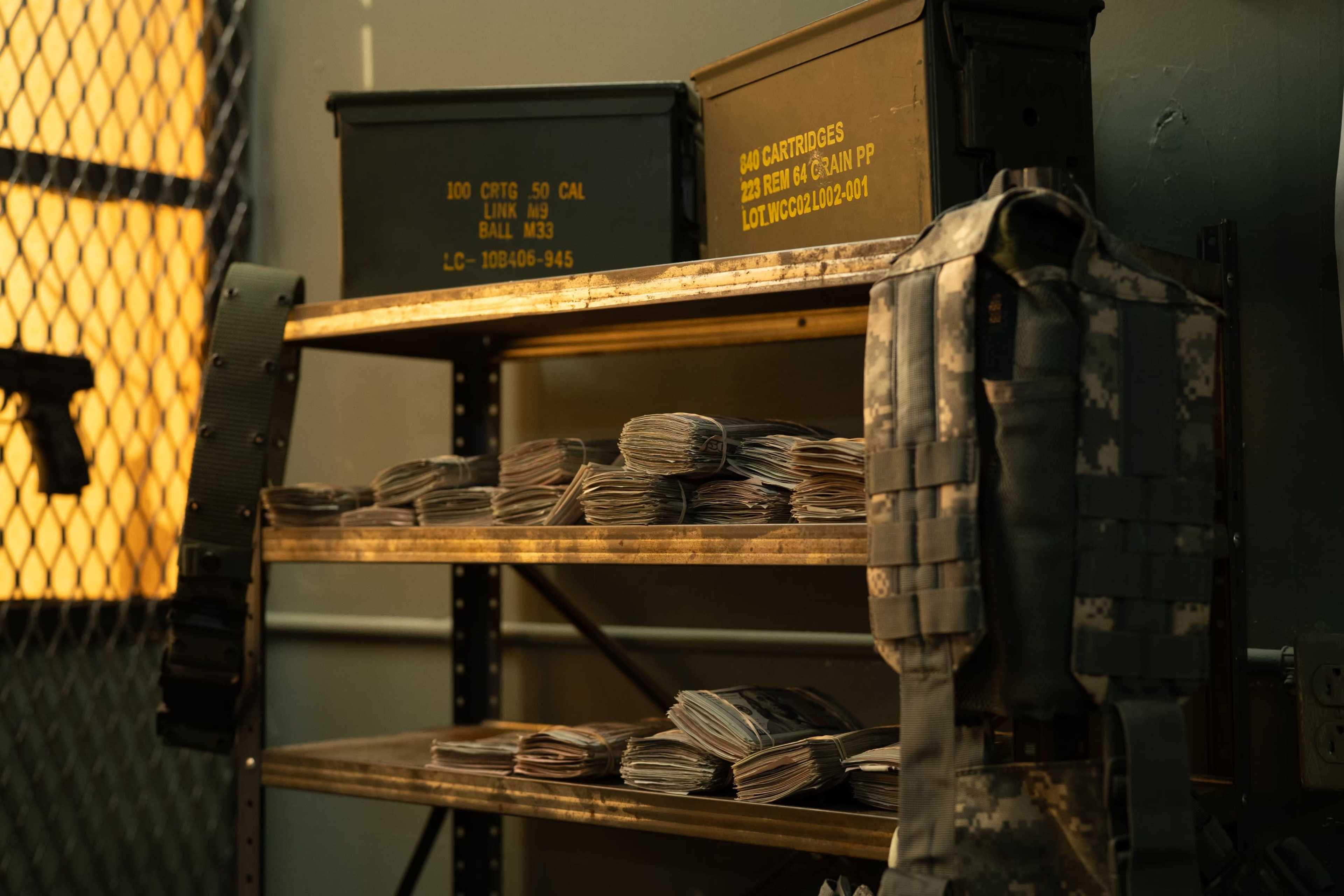 A shelving unit holds stacks of cash alongside military gear and fortified containers in a dimly lit room.