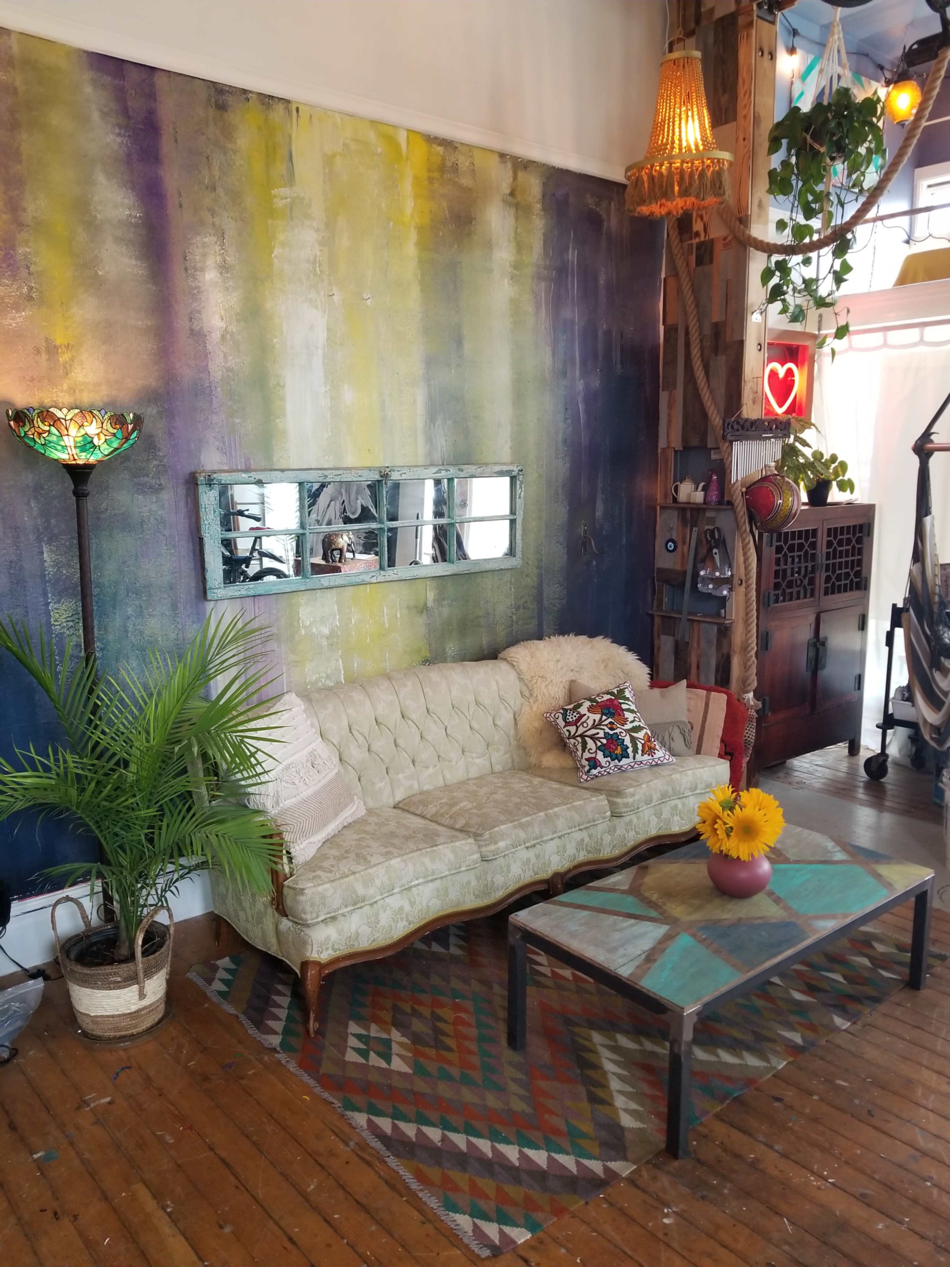 A vibrant living room with a green tufted couch, a patterned rug, a coffee table with a vase of sunflowers, and a colorful wall adorned with plants and decor.