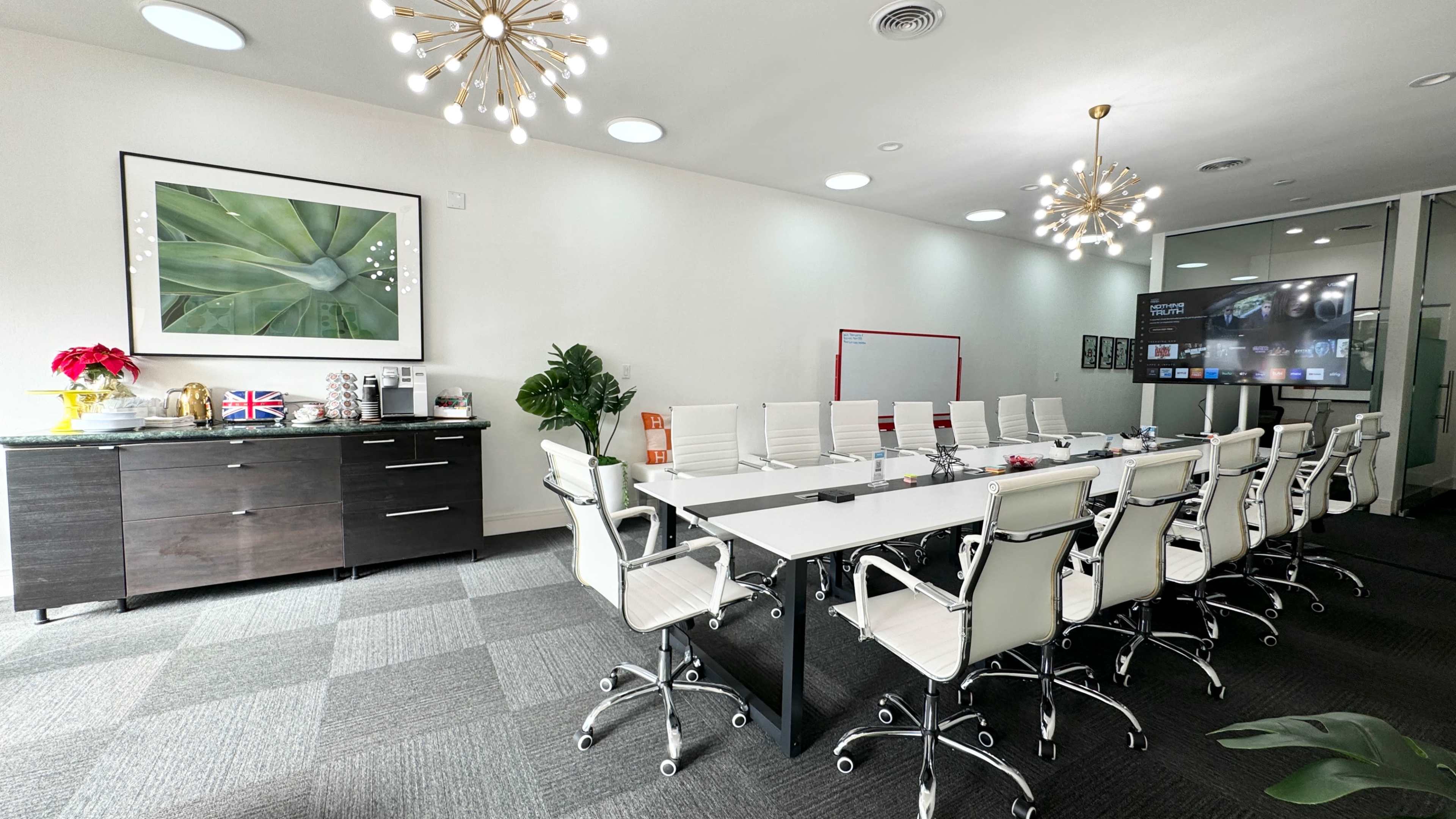 A modern conference room features a long table surrounded by white chairs, a large screen on one wall, and decorative elements including plants and a framed picture.