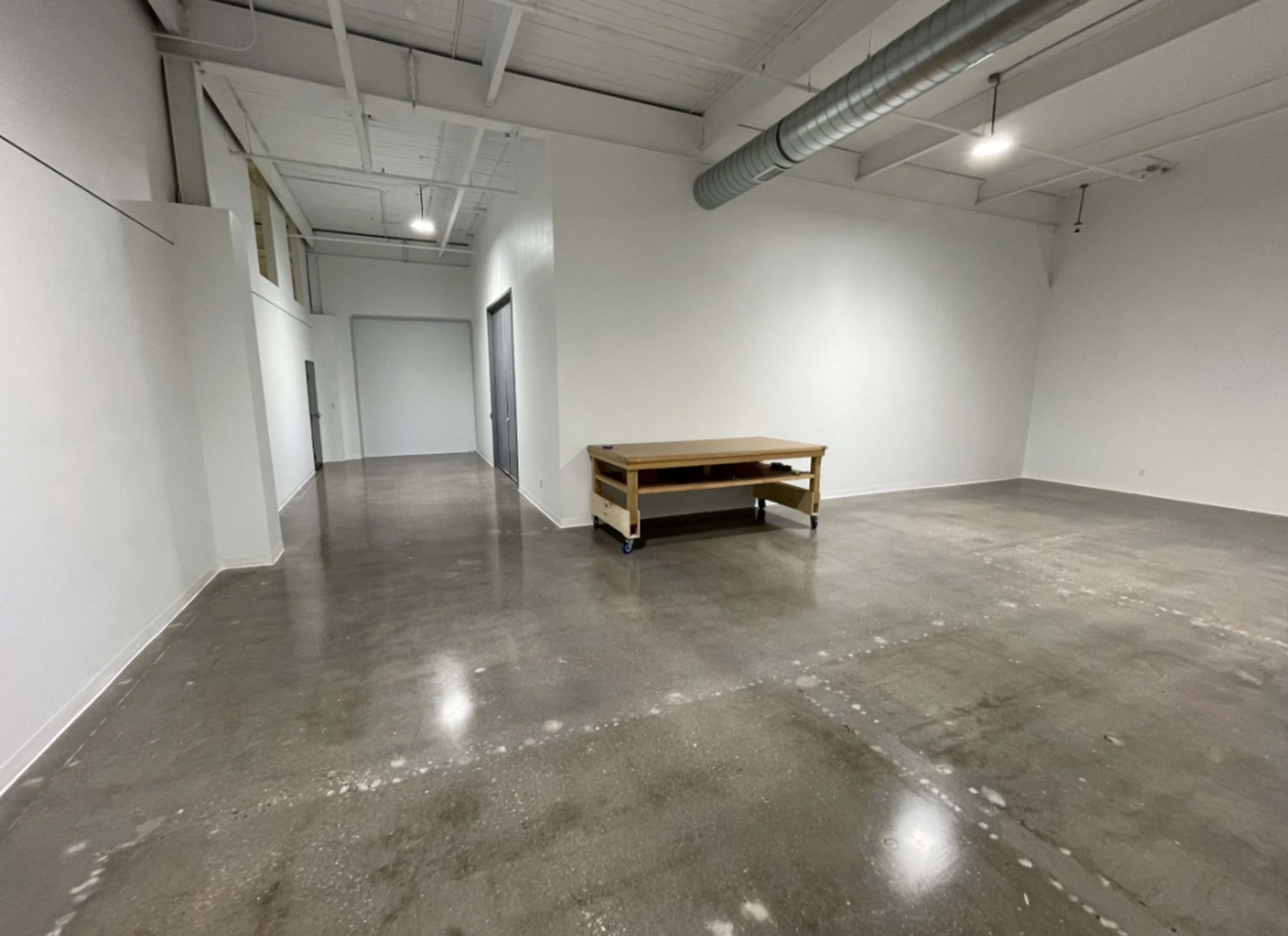 An empty industrial space with concrete flooring, white walls, and a wooden table in the center.