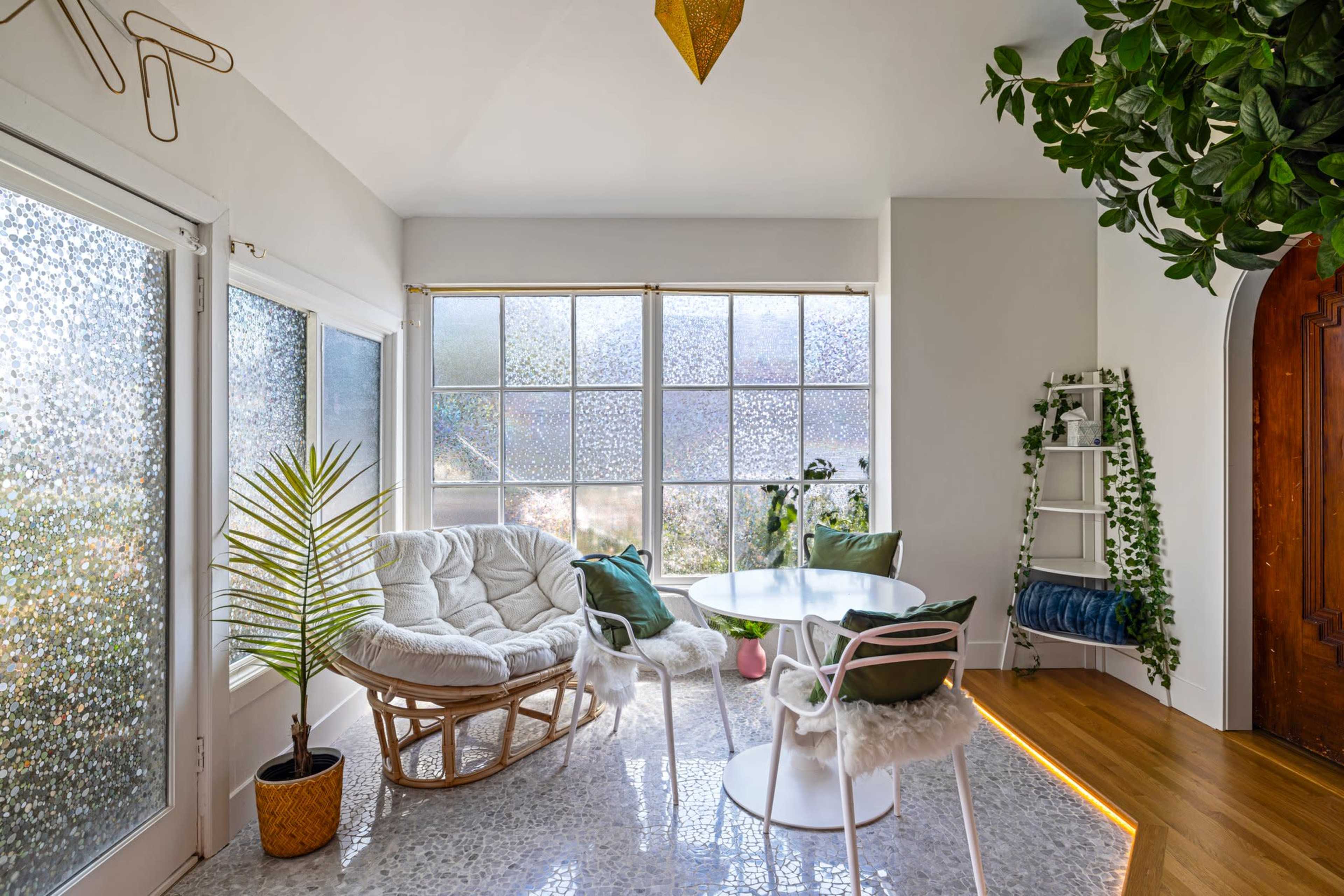 A bright room features large windows with a textured glass design, a cozy white seating area, a round dining table, and greenery along the walls.
