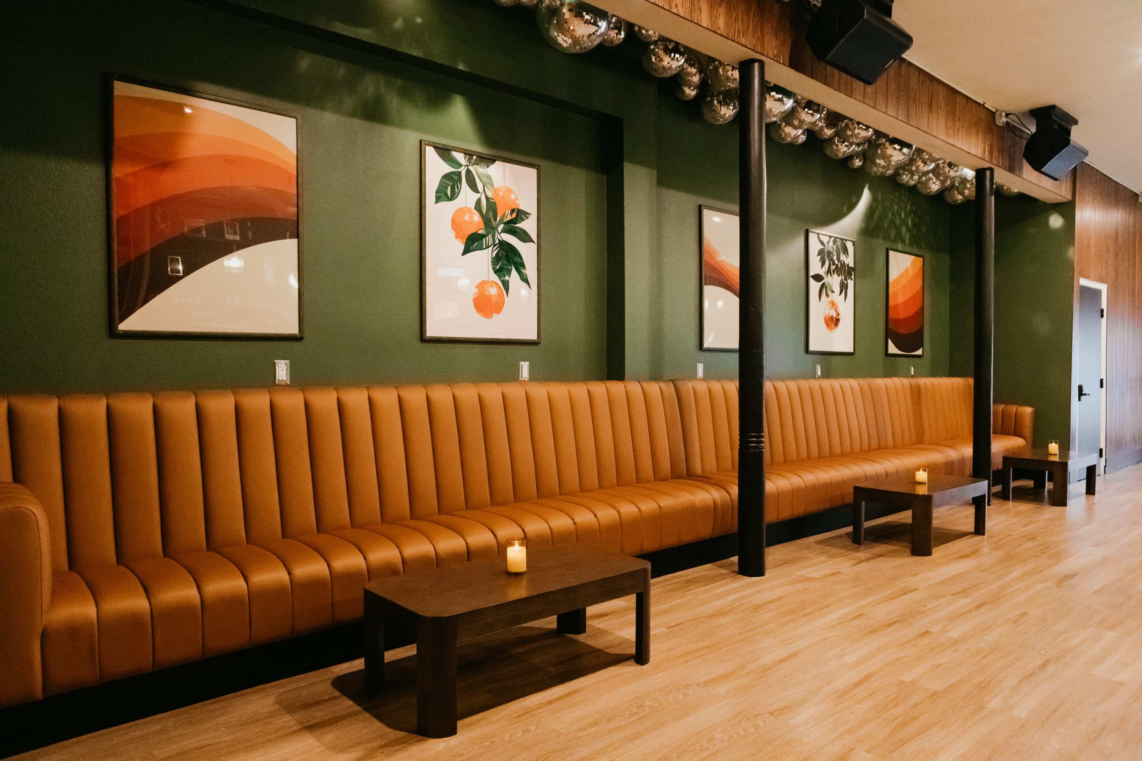 The image shows a modern lounge with a long, brown leather banquette against a green wall, featuring framed artwork and small tables with candles.