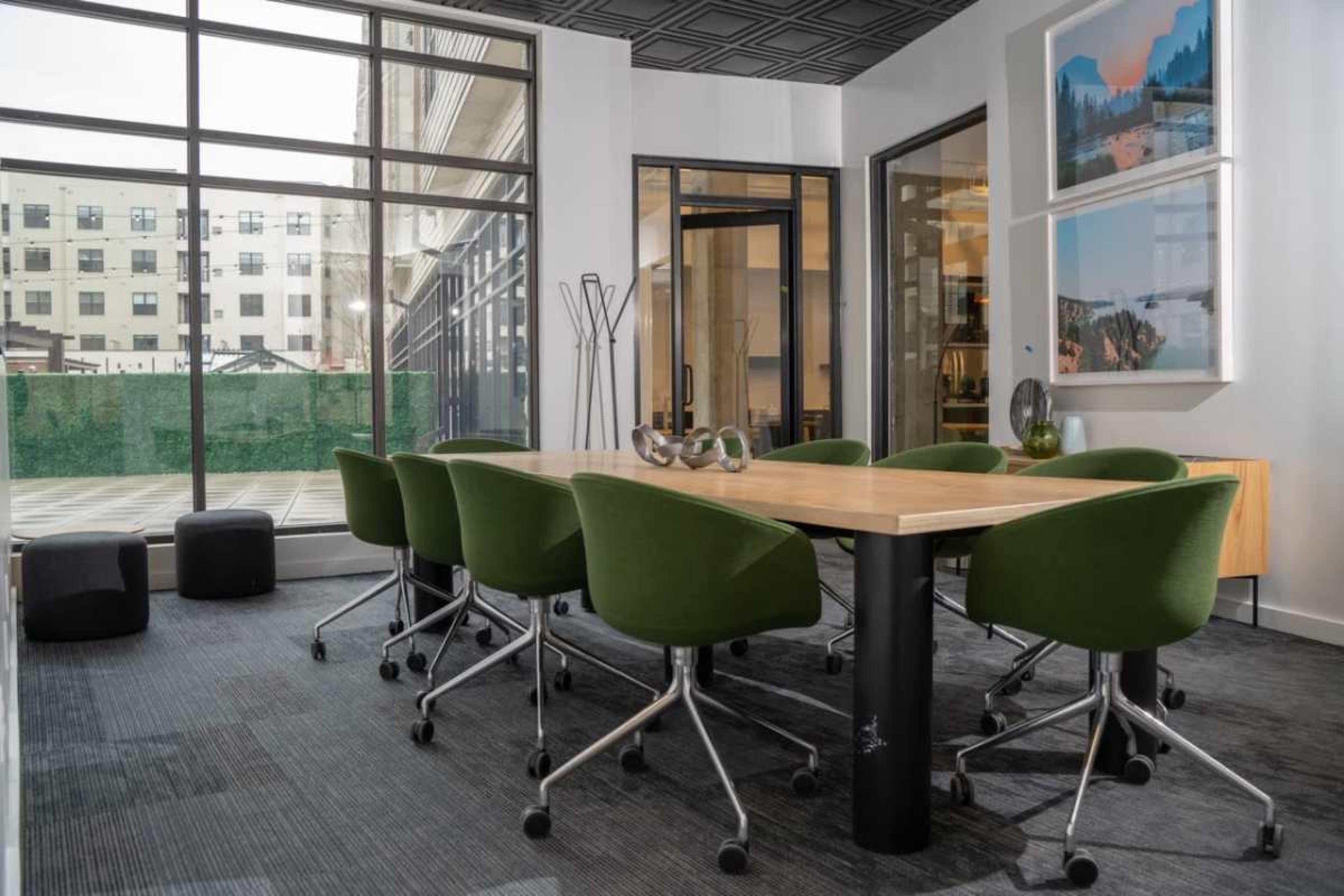 A modern meeting room features a wooden table surrounded by green chairs and large windows that provide a view of an outdoor space.