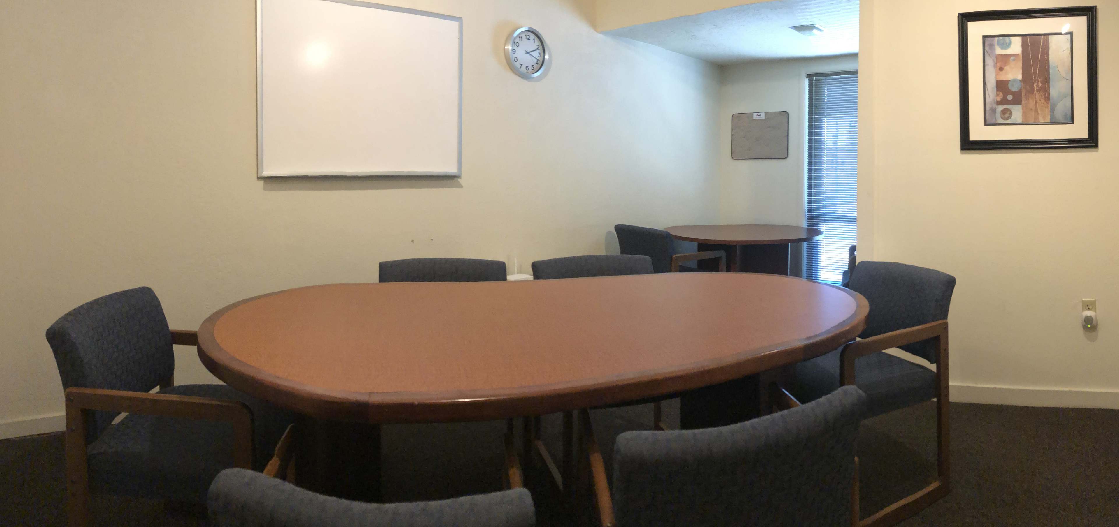The image shows a small conference room with an oval table surrounded by eight chairs, a whiteboard on the wall, and a clock.