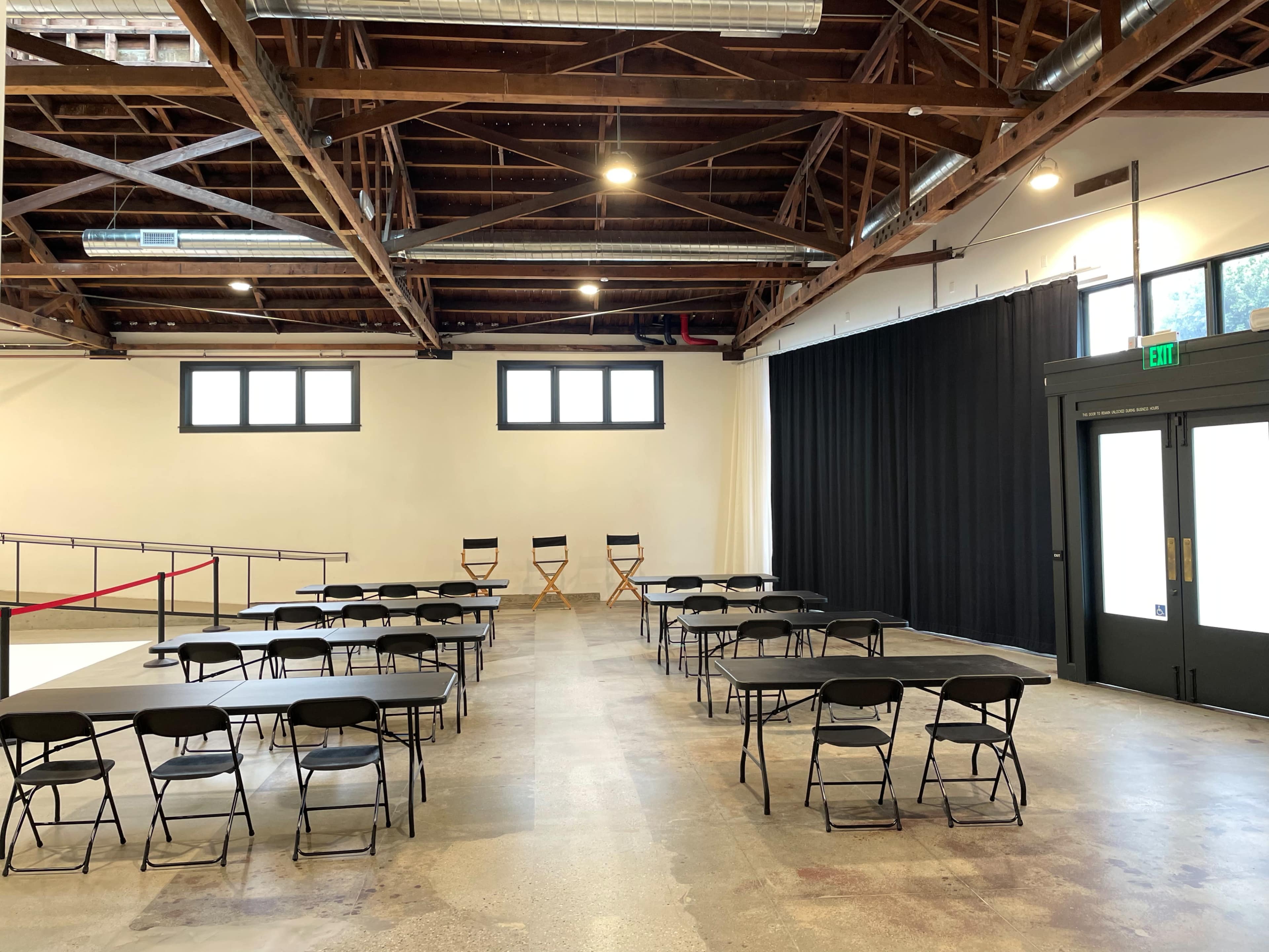 The image shows a spacious room with several rows of black folding tables and chairs arranged neatly, along with a black curtain and two director's chairs positioned against the wall.