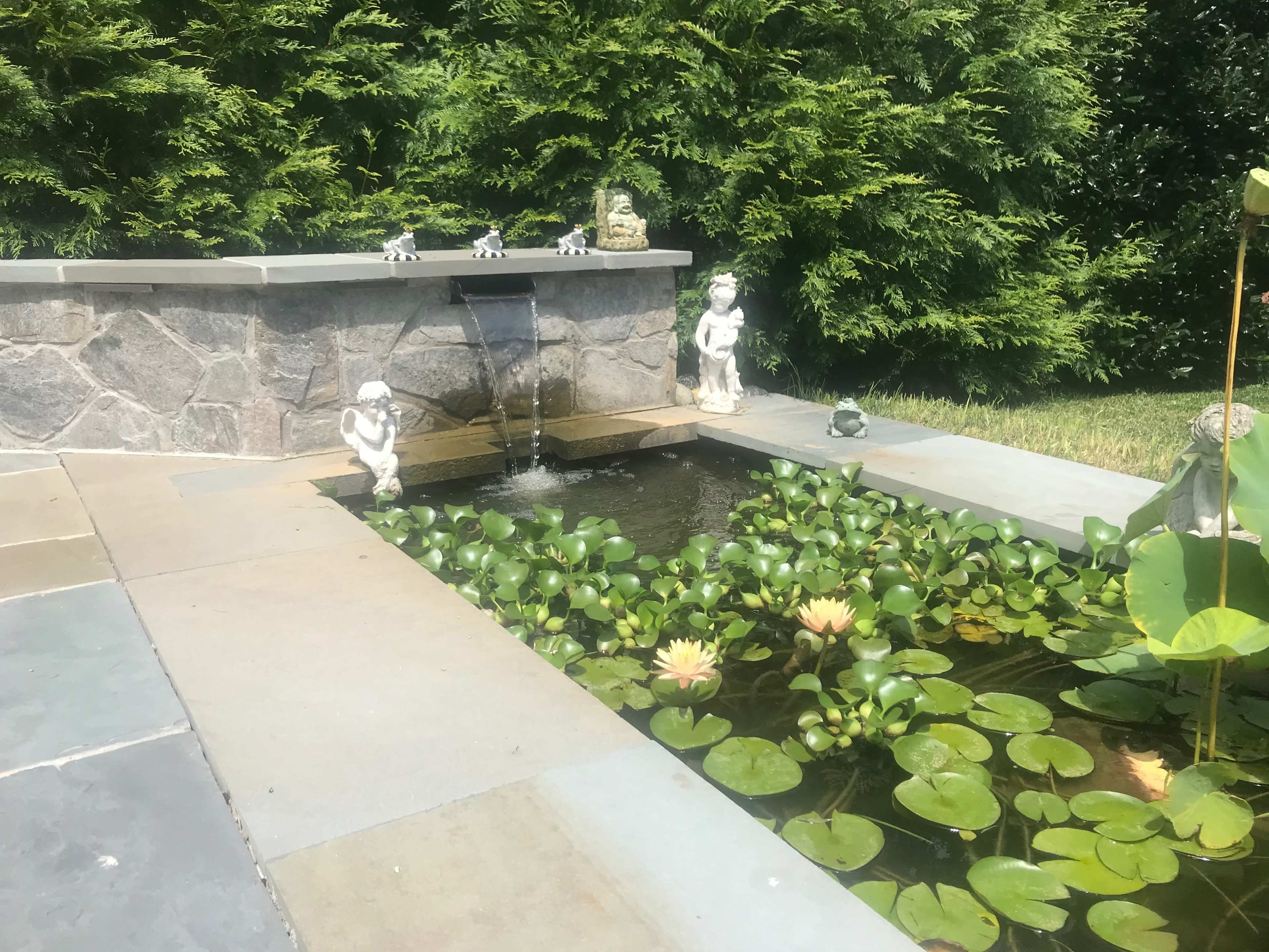 A stone pond with a small waterfall features statues of children on either side and water lilies on the surface.