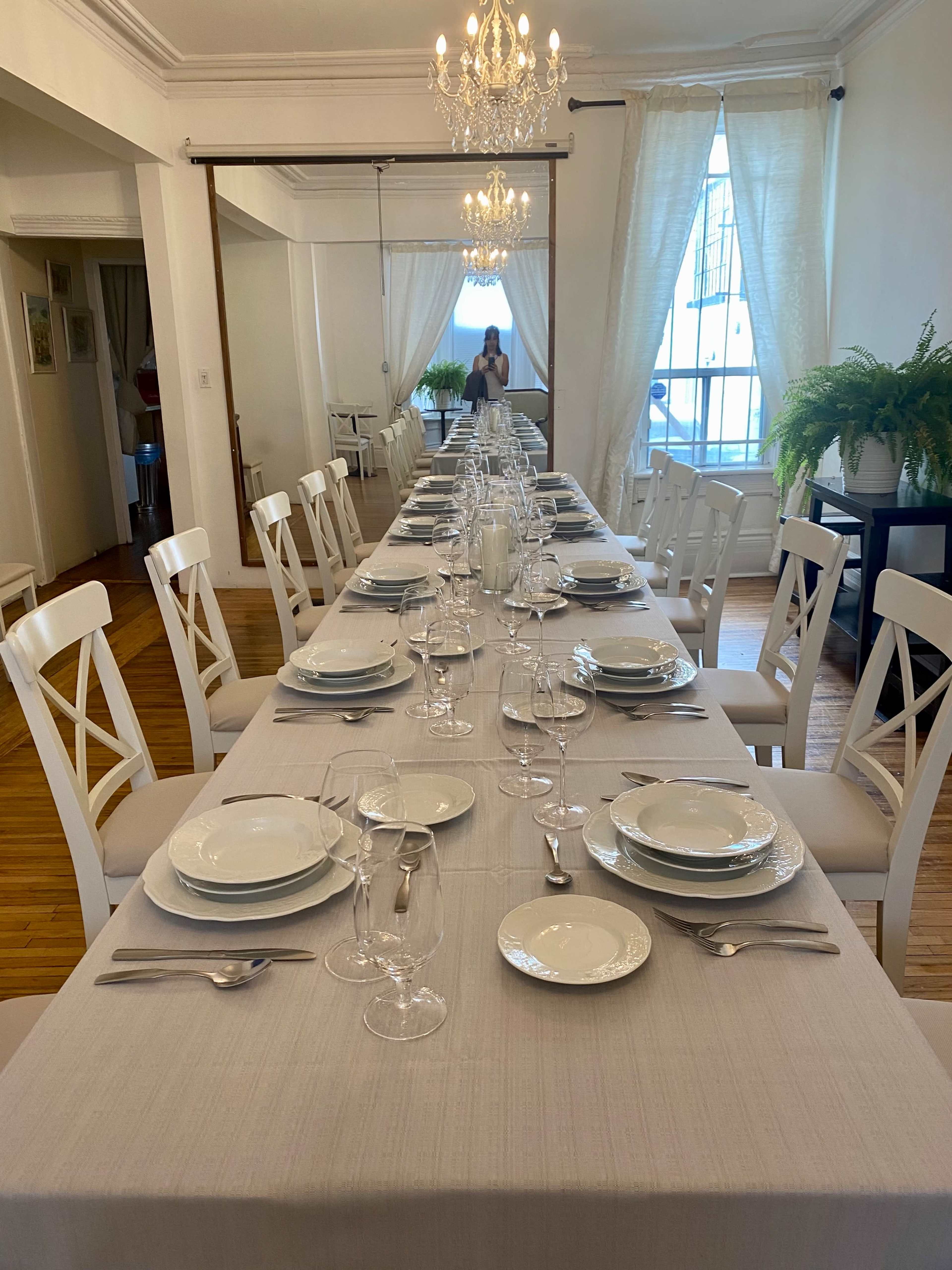 A long dining table is set with plates, glasses, and cutlery under a chandelier in a well-lit room.