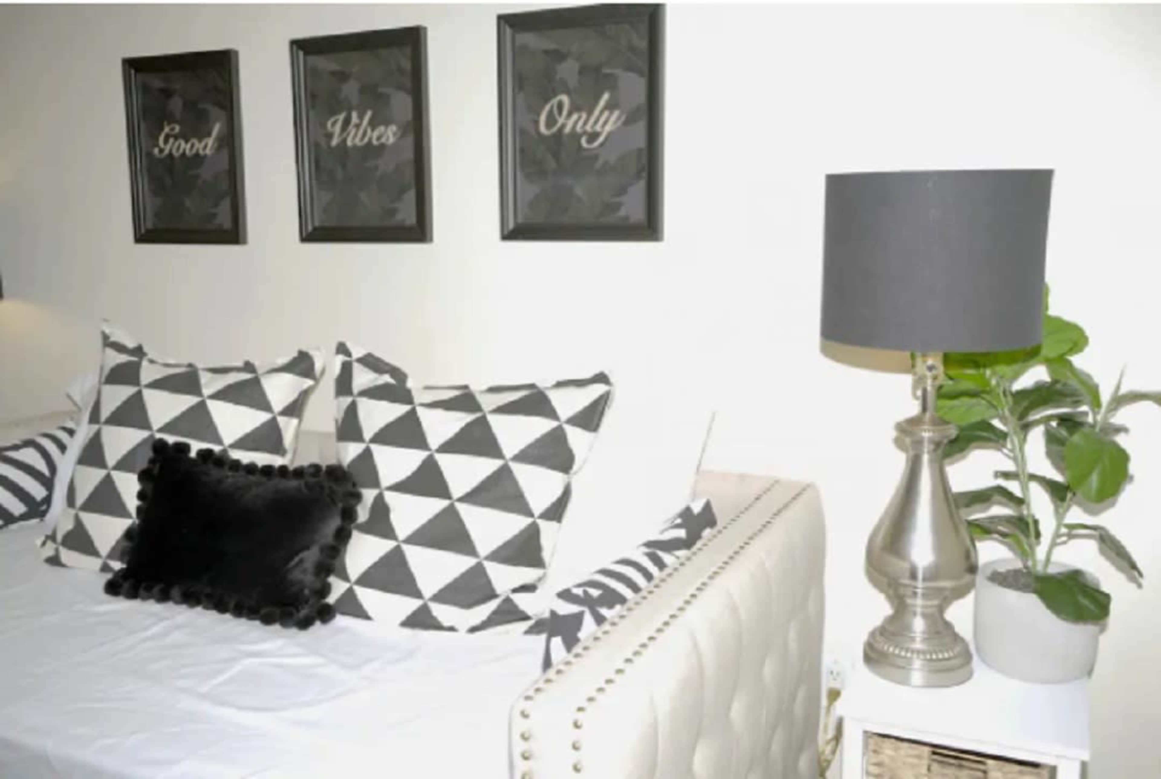 The image shows a cozy interior featuring a bed with patterned pillows, three framed wall decorations, a lamp, and a potted plant.